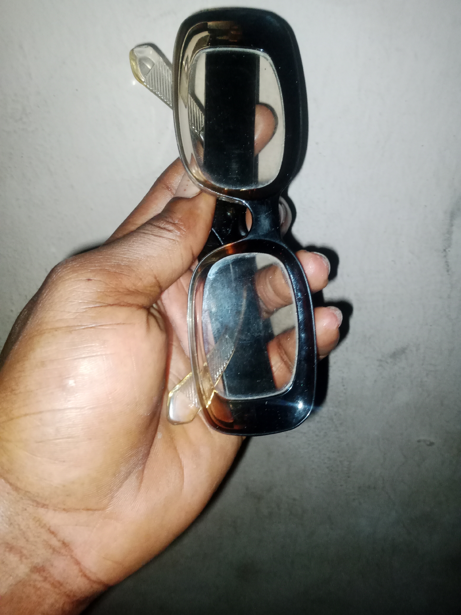 A person's hand is holding a pair of eyeglasses against a light gray, textured wall in Jauro Yinu, Nigeria. The eyeglasses have dark frames and clear lenses. The hand is dark-skinned and appears to be an adult's. The lighting is dim, creating shadows on the wall.