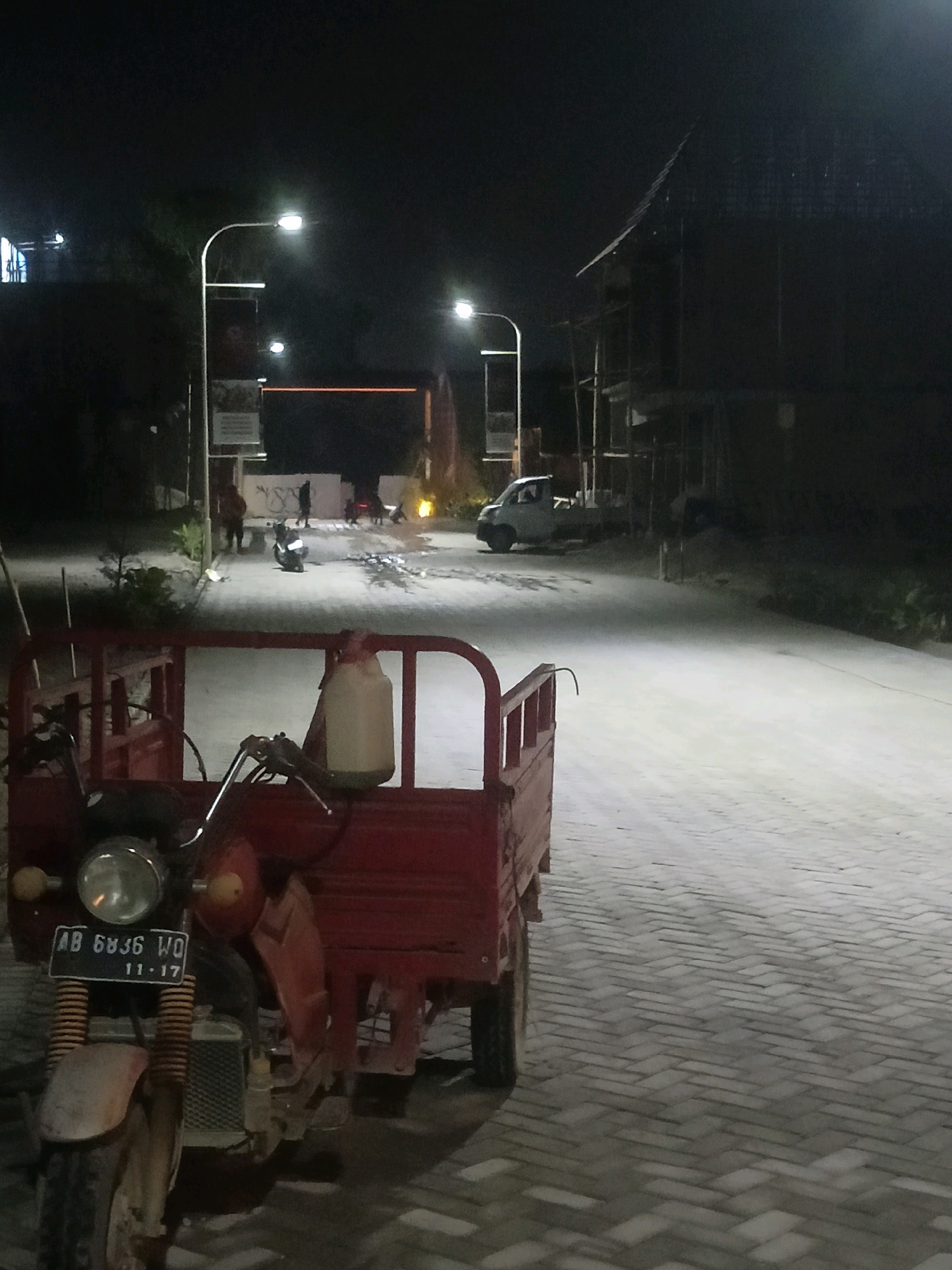 At night, a red three-wheeled motorcycle with a cargo bed is parked on a paved road in Sendangadi, Indonesia. The motorcycle has a visible license plate reading "AB 6836 WO 11-17". Streetlights illuminate the scene, casting long shadows. In the background, a few individuals are standing near other parked motorcycles and a small white truck. A building structure, possibly under construction, is visible to the right, with scaffolding and an angled roof. The ground is paved with interlocking bricks, and some scattered debris is present on the road.