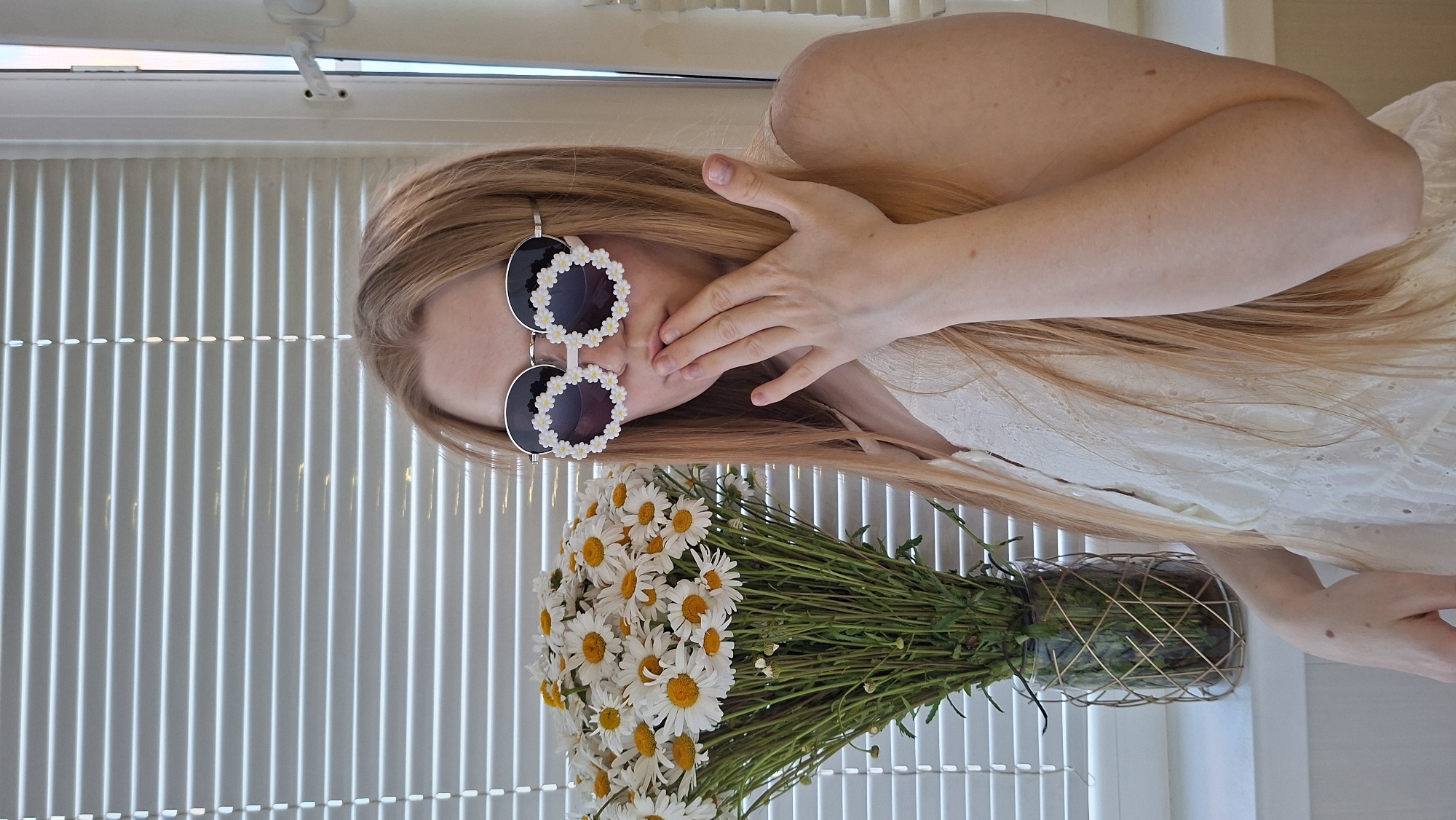 A young woman with long, light brown hair is posing for a photo indoors. She is wearing a white eyelet dress and oversized, round sunglasses adorned with daisy shapes around the frames. She is covering her mouth with her right hand, with her fingers extended. To her left, a large bouquet of white daisies with yellow centers is in a clear glass vase with a gold wire cage. The background consists of a window with horizontal blinds and a white windowsill. The image appears to have been taken in Chaikovsky, Russia.