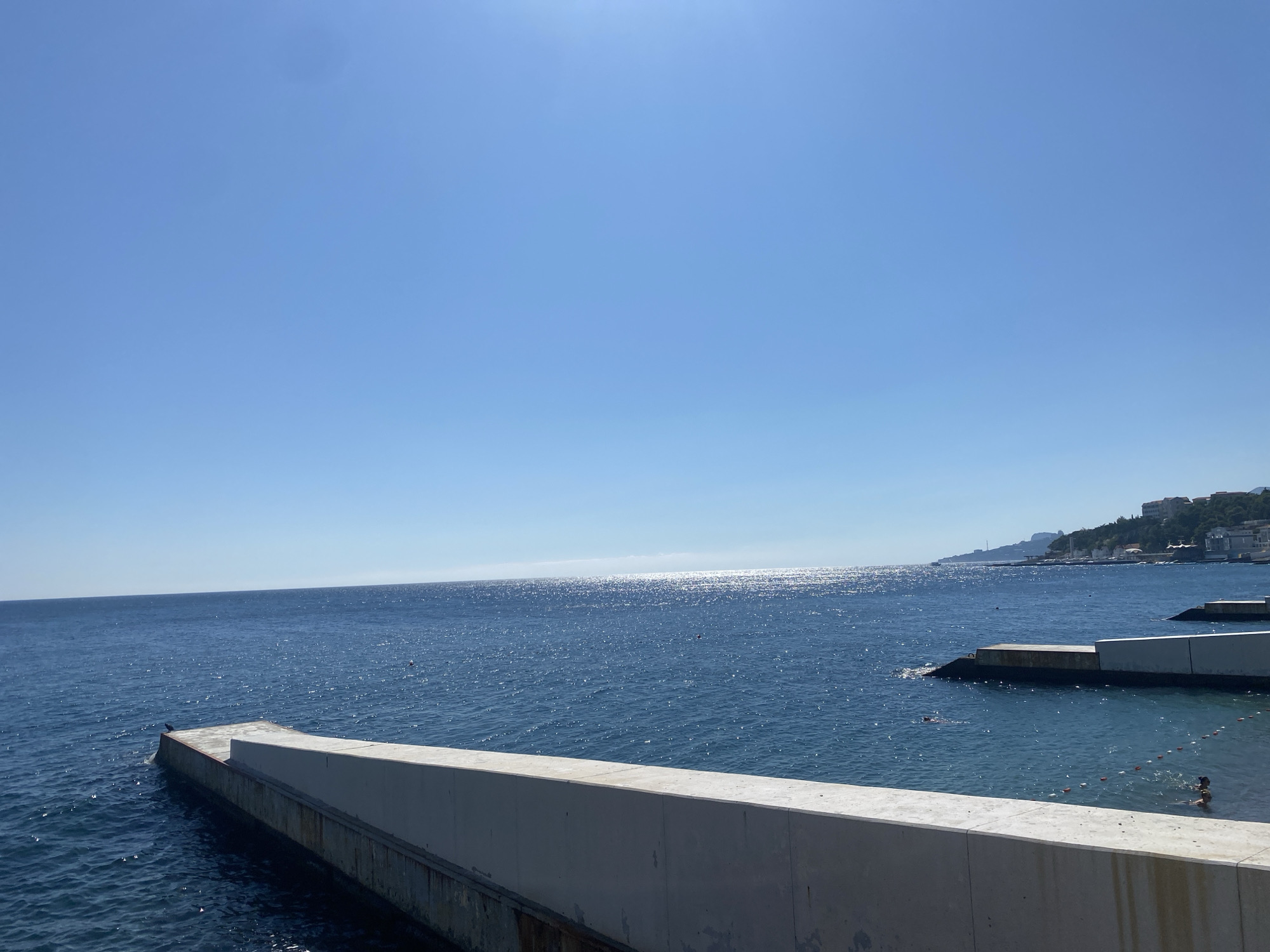 The image shows a clear blue sky over a tranquil blue sea. In the foreground, a concrete structure, possibly a pier or seawall, extends into the water. A small black bird is perched on the edge of the pier. Further out in the sea, several people are swimming. In the distance, a coastline with buildings and greenery is visible under the bright sun. The overall scene suggests a peaceful day at the seaside in Гаспринский поселковый совет, Ukraine.