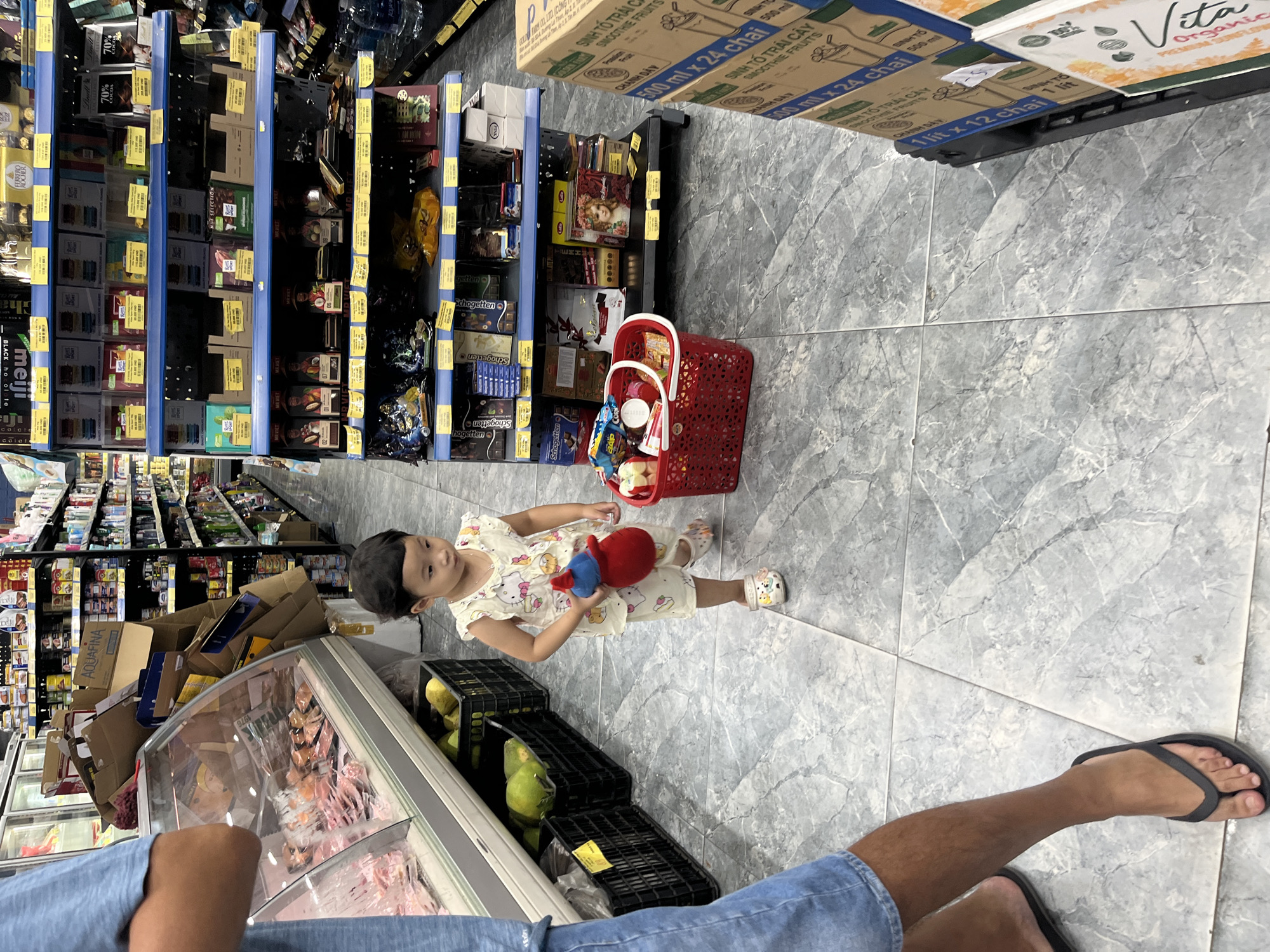 The image shows the interior of a grocery store, possibly in the Bac Nha Trang Ward in Vietnam, based on the OCR text "SINH TỐ TRÁI CÂY," which is a Vietnamese phrase for "fruit smoothie." A young child, dressed in a light-colored patterned outfit and sandals, stands in the aisle holding a red and blue toy. A red plastic shopping basket sits beside her. The store shelves are stocked with various products, including boxes of chocolate, and beverages. In the foreground, an adult wearing a light blue shirt, shorts, and flip-flops stands near a refrigerated display case containing various food packages. Several crates containing produce, possibly coconuts, are on the floor. The floor is covered in grey-white tiles.