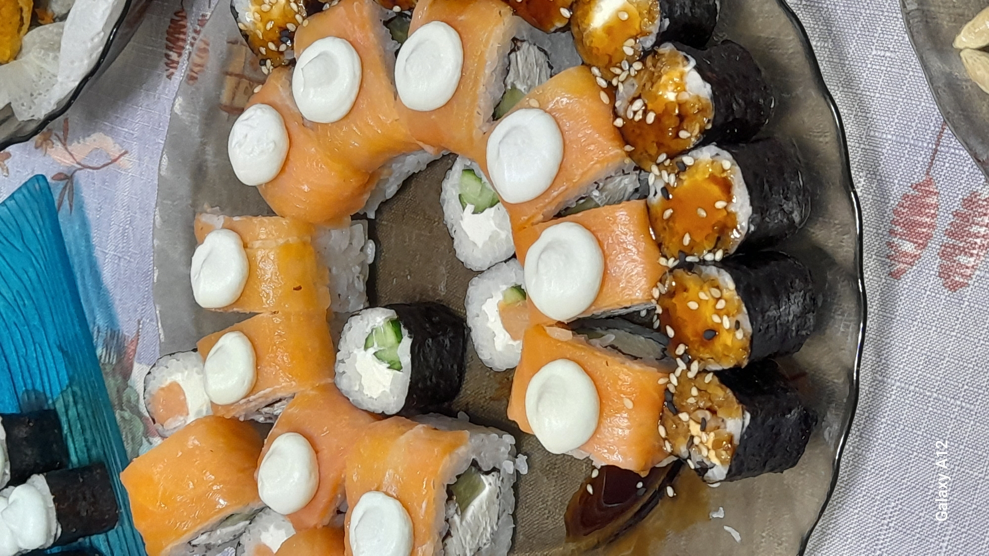 The image displays a close-up overhead view of a sushi platter, captured indoors, presumably at a dining table in Марковский, Russia. The central subject is a clear, irregularly shaped serving plate laden with assorted sushi rolls.
Multiple types of sushi are present:
1. **Salmon-wrapped rolls**: Numerous rolls are covered with thin slices of orange salmon, revealing white rice, cream cheese, and green cucumber fillings. Many of these rolls are topped with distinct dollops of white cream, likely cream cheese or mayonnaise.
2. **Nori-wrapped rolls**:
* Several dark green/black nori-wrapped maki rolls are visible, primarily featuring white rice and green cucumber within. One such roll is prominent in the upper-center.
* Other nori-wrapped rolls are topped with a glossy, light brown sauce, speckled with white and black sesame seeds. The visible filling under the sauce appears light brown/orange.
3. **Other rolls**: Some rolls in the upper background are partially visible, consistent with the salmon-wrapped style.
The plate rests on a light-colored, possibly grey or beige, woven tablecloth with a subtle texture and a faint, light red/pink leaf-like pattern in the lower right corner. The background is partially blurred, showing indistinct shapes including a blue, textured object in the upper center, and what appears to be part of a bowl with light contents to the upper right. Further right, a yellow/orange fried item is partially visible. Out-of-focus light-colored oval objects, possibly nuts or seeds, are present in the lower right background. The watermark "Galaxy A12" is visible in the bottom left corner. No individuals are depicted. The scene suggests a prepared meal or dining setting.
