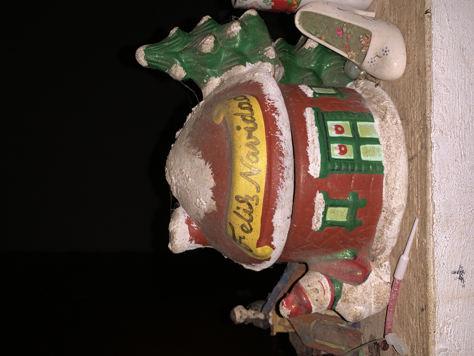 An upward-facing photographic image displays a collection of holiday-themed figurines situated on a light-colored, textured surface in Santa Ana, Venezuela. The central subject is a rounded, red ceramic house, approximately 15-20 cm in height, with a prominent white, textured material simulating snow on its roof and chimney. A yellow banner affixed to the roof displays "Feliz Navidad" in yellow script. The house features green-framed windows with yellow panes, some adorned with small red dots.
To the immediate right of the house, a green ceramic Christmas tree, also topped with simulated snow and exhibiting fine cobwebs, stands upright. To the left, a smaller snowman figurine, wearing a red hat and green scarf, is positioned. Further to the left and in the background, a small, partially obscured figurine of a person, possibly a male, dressed in dark attire, is visible.
In the lower right foreground, a miniature white high-heeled shoe figurine, decorated with blue floral patterns, contains green fibrous material and small pink decorative elements within its opening. In the lower-center foreground, a pink glitter-coated rod with a white plastic cap on one end and a pointed white tip on the other lies on the surface. The background is uniformly dark, suggesting low light conditions or a black backdrop, with primary illumination directed at the figurines. The figurines and the surface exhibit visible dust and minor wear, consistent with aged decorative items.