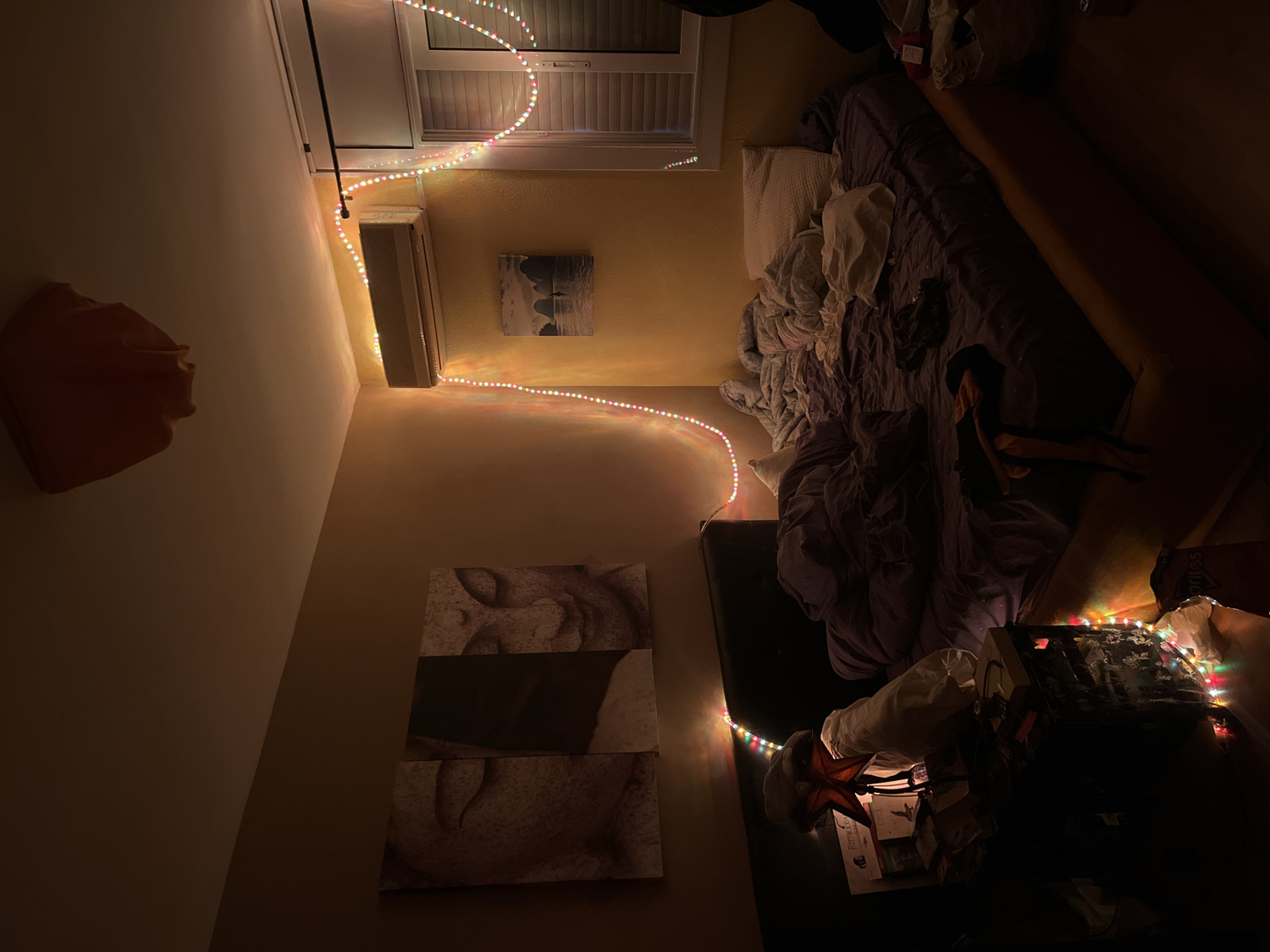 The image displays a dimly lit, cozy room, likely a bedroom or living area, viewed from a high angle looking down. The primary illumination comes from colorful LED string lights tracing the walls and furniture, creating a warm, relaxed atmosphere, suggesting it is nighttime or late evening.
On the left side of the room, a light-colored wall features an abstract, reddish-brown, organic-shaped sculpture mounted high up. Below it, three rectangular panels of abstract, textured art are stacked vertically. The far wall, painted a light yellow or beige, has an air conditioning unit installed high, beneath a window fitted with white blinds or shutters. A framed picture, possibly a landscape or seascape, hangs below the AC unit.
The multi-colored LED string lights run along the ceiling line, descend the far wall past the air conditioner, and continue along the edge of what appears to be a bed or a partition, extending into the foreground.
On the right side of the image, a large bed is visible with rumpled purple and grey bedding and several pillows. A person is lying face down or turned away on the bed, partially covered by a blanket. Adjacent to the bed, along the right edge, is a long, padded surface, possibly a couch or an extension of the bed, in a warm brown tone. Another person's legs are visible, resting on this surface.
In the bottom right corner, a nightstand or small table is cluttered with various items. A lamp with a star-shaped shade sits among books, one of which clearly displays "Hermes" and potentially "Fictions" on its spine. More colorful string lights are draped over what appears to be a media console or similar electronic device on the table. The room has a lived-in feel, with scattered items contributing to a casual, comfortable ambiance. The location context provided is Barcelona, Spain.