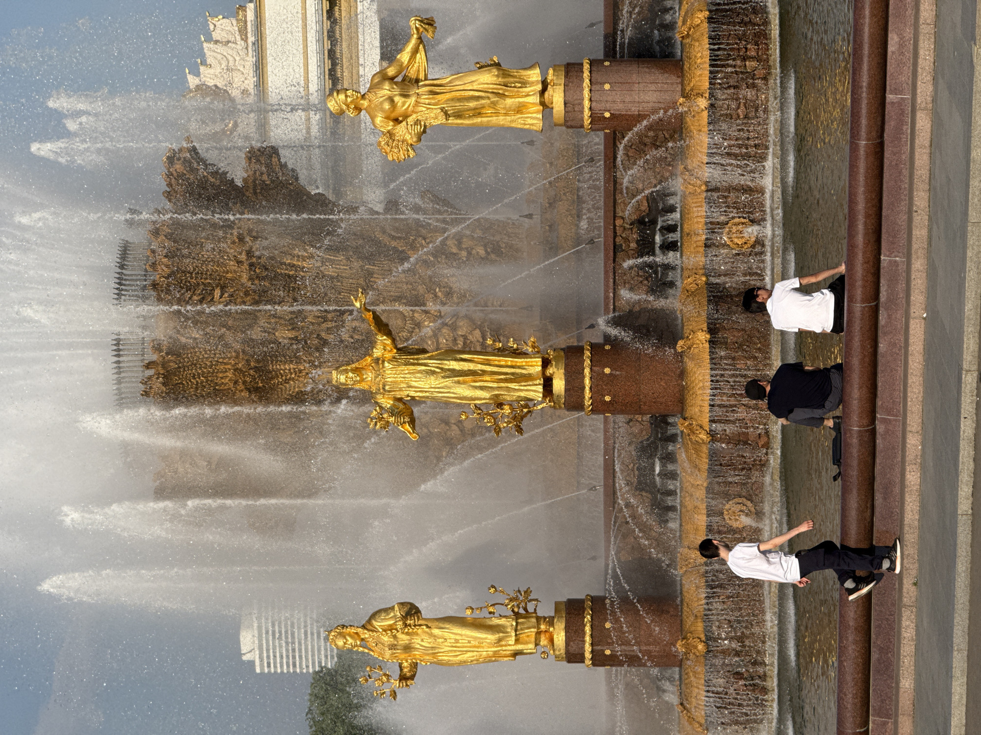The image shows the Friendship of Nations Fountain in Moscow, Russia. The fountain features several golden statues of women on pedestals, representing different Soviet republics. Water cascades from the fountain in multiple tiers. Three people are visible alongside the fountain's edge. One person is wearing a white shirt and dark pants, another person is wearing a black shirt and dark pants, and the third person is wearing a white shirt and dark pants. The architecture of a building is visible in the background.
