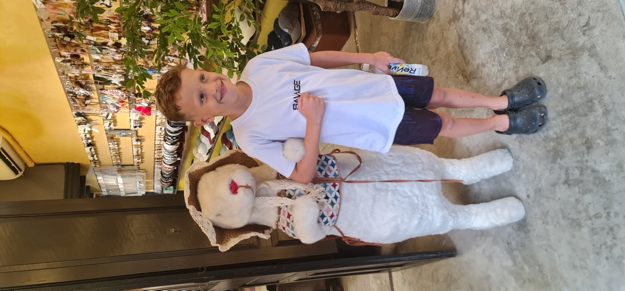 A portrait shot captures a fair-skinned boy with short, light brown hair, clad in a white "SAVAGE" t-shirt and dark shorts, holding a "Revive" bottle, standing alongside a large stuffed animal resembling a white bear wearing a bonnet and a saddlebag with a patterned design. The foreground features a gray concrete floor, while the background reveals a shop interior with shelves filled with various items, including hats and headbands, under a yellow wall and a green, leafy plant. The setting is in Hội An Ward, Vietnam.
