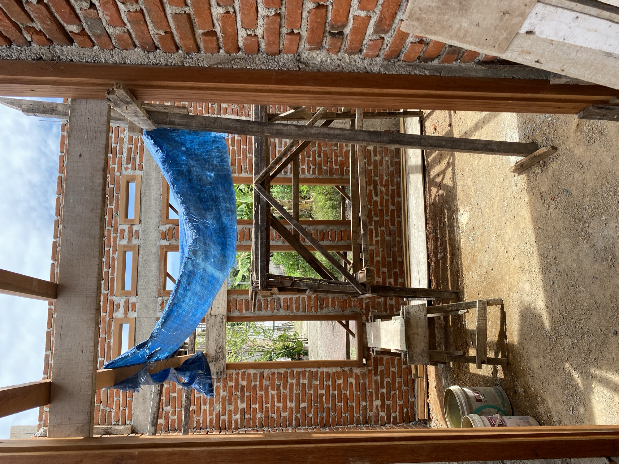 The image depicts the interior of a building under construction in Banda Aceh, Indonesia. The structure is made of red brick walls and features exposed wooden beams and supports for the roof and frame. A blue tarp is draped over some of the wooden scaffolding, suggesting it's being used for protection from the elements. There are several window openings in the brick walls, some with wooden frames already installed. The ground inside the building is dirt and gravel. In the foreground, to the left, are two buckets, one white and one light green. To the right, leaning against the wall, is a piece of plywood. Scaffolding made of wood is visible in the center and right of the frame. The overall scene is one of ongoing construction work.