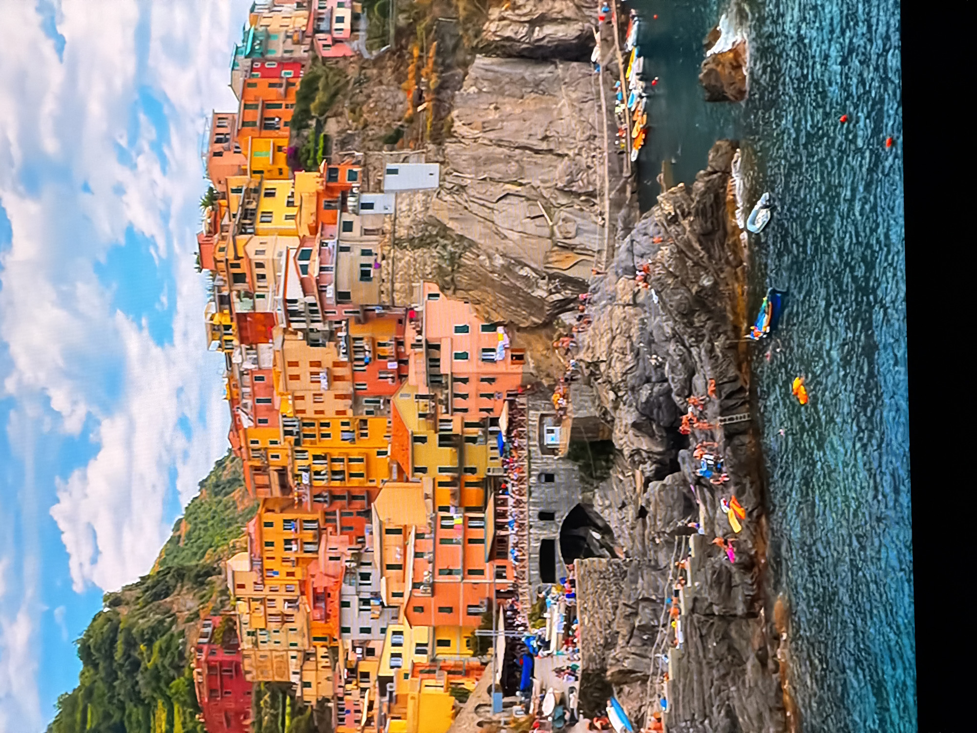 The image depicts a vibrant coastal town with densely packed, colorful buildings cascading down a rocky hillside towards the sea. The architecture is characterized by multi-story structures painted in hues of orange, yellow, pink, and red, with visible windows and balconies. The town is situated on a steep, uneven terrain, with some buildings built directly into the rock face.
The foreground shows a rocky shoreline with people sunbathing and relaxing on the stones. A few small boats are moored in the clear blue water. Further back, a paved area or walkway runs along the base of the cliffs, populated by more people and lined with small boats. A large stone archway is visible, leading into the town.
The sea is a deep blue-green, with small waves and ripples on the surface. Several boats, including small motorboats and rowboats, are scattered in the water and along the harbor area. Some are brightly colored.
The sky is a bright blue with scattered white clouds. The overall scene conveys a sense of a bustling, picturesque Mediterranean or Italian coastal village, likely a popular tourist destination.
The city of Chirchik, Uzbekistan, is not depicted in this image. The visual elements are inconsistent with the geography and architecture typically associated with Chirchik, which is an inland city in a more arid region and does not have a coastal setting.