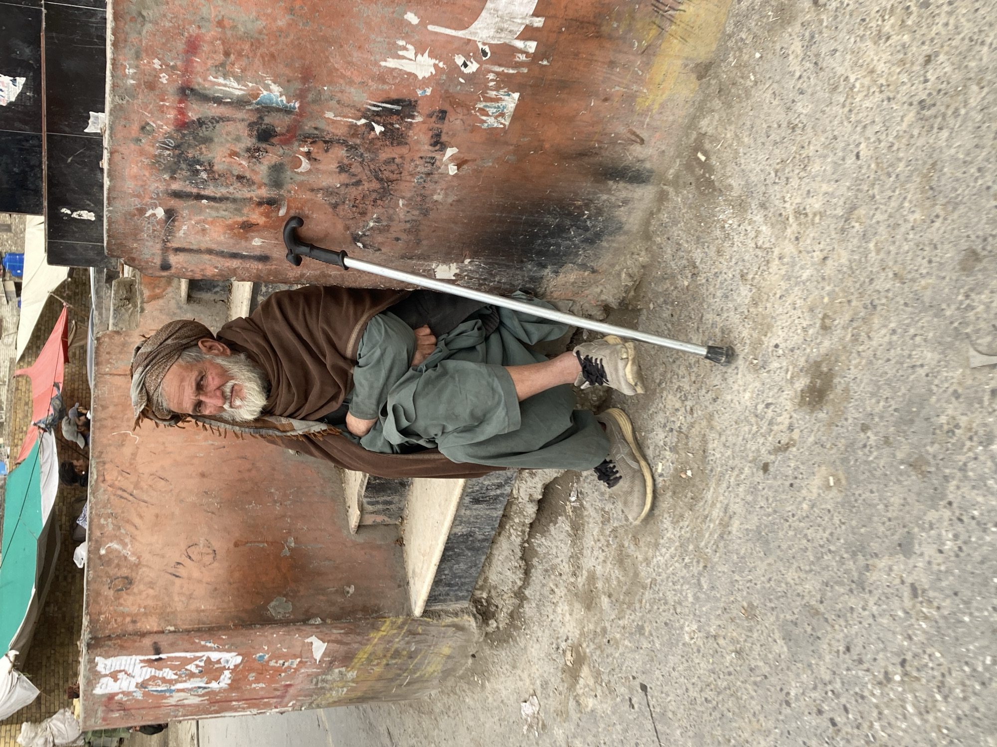 An elderly man is seated on the second step of a concrete staircase or platform in an outdoor urban environment within Quetta City Tehsil, Pakistan. He has a long white beard and mustache, wears a light brown turban, a dark brown shawl draped over his shoulders, and teal or olive green traditional attire with light grey sneakers. His arms are crossed, and his hands are tucked into his clothing. A silver walking cane with a black handle rests upright against the ochre-colored concrete wall to his right. This wall exhibits extensive wear, various graffiti markings, and remnants of torn paper or posters. The ground in the foreground is rough, grey concrete. In the background, behind the wall and to the left, are multiple makeshift shelters constructed from white, green, and red tarpaulins, with several indistinct individuals visible, one wearing a grey cap. Distant buildings, including some with blue rooftop water tanks, are present in the far background. The man gazes directly forward.