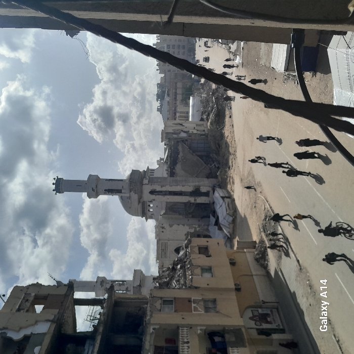 The image captures a scene of extensive urban damage, likely in a conflict-affected area, viewed from an elevated perspective. The sky above is partly cloudy with bright daylight.
In the mid-ground, a prominent mosque with a tall, slender minaret and a dome stands out, though it exhibits visible signs of damage, including chipped surfaces and possibly structural compromise. Around the mosque and extending into the foreground, numerous buildings are in various states of disrepair and destruction. Many structures appear to be shelled or collapsed, with rubble scattered on the ground and exposed interiors, rebar, and skeletal frameworks visible.
A wide, paved street runs through the scene, stretching from the mid-ground towards the background. Several human figures are present on this street, primarily identifiable by their distinct, elongated shadows cast by the sun. These shadows suggest people are walking, and one shadow appears to belong to someone riding a bicycle. The actual individuals casting these shadows are small and less distinct in the distance.
The long shadows on the street indicate that the sun is relatively low in the sky, suggesting either mid-morning or late afternoon. The clouds are scattered and appear white and grey against a lighter blue sky.
In the immediate foreground, a thick, dark cable or pipe runs diagonally across the upper portion of the frame, partially obscuring the view of the sky and some distant buildings.
A watermark with the text "Galaxy A14" is visible in the bottom right corner of the image. The overall impression is one of a city grappling with widespread destruction, yet still seeing signs of life with people moving through its damaged streets.