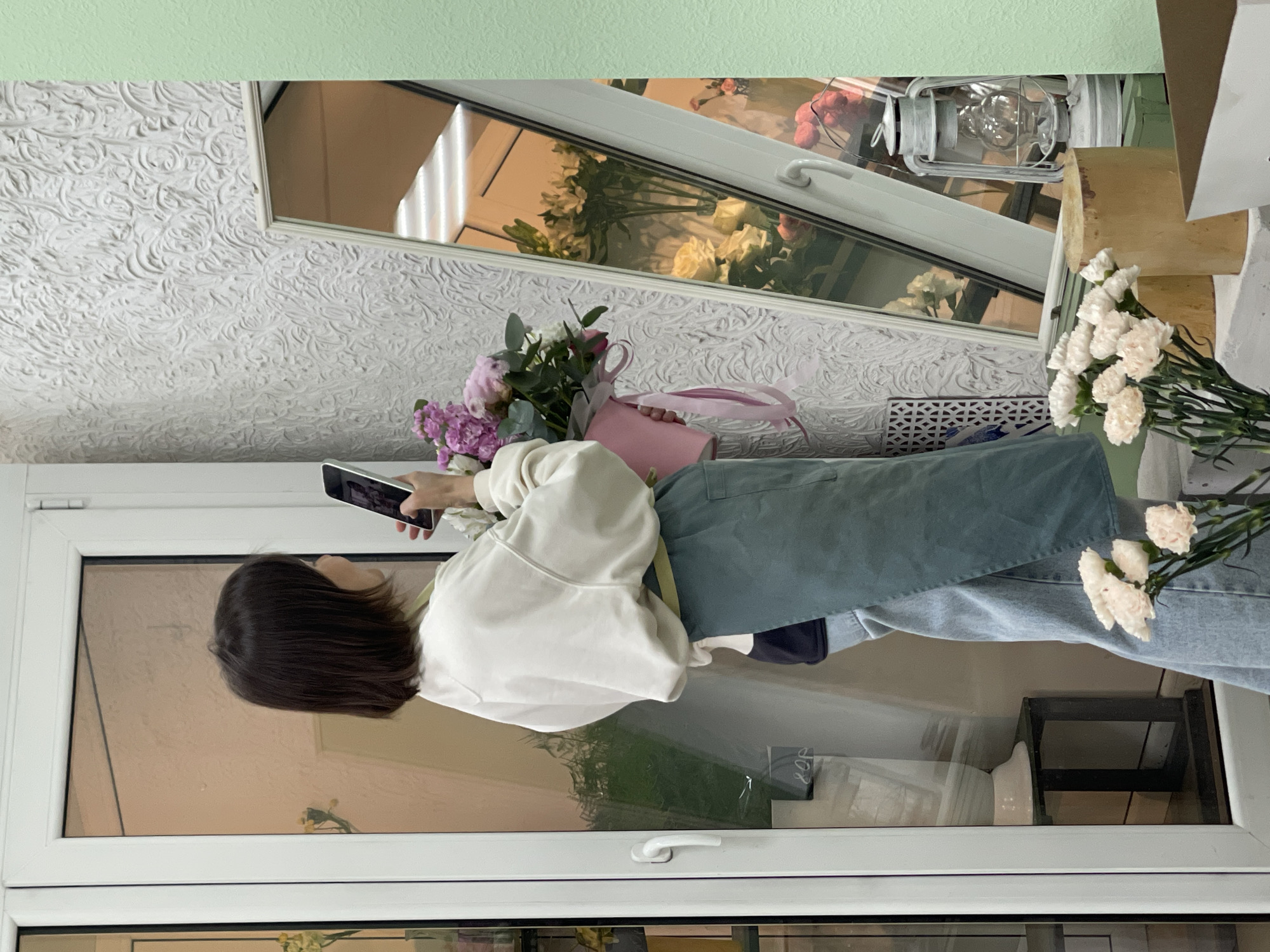 A woman with short dark hair is taking a picture with her phone of a bouquet of flowers in a pink bag. She is wearing a white sweater and blue pants. The scene appears to be inside a building, possibly a store or a residence, with a window reflecting the outside. There are other flowers visible in the background and a decorative lantern. The location in the city is Unknown, Unknown.