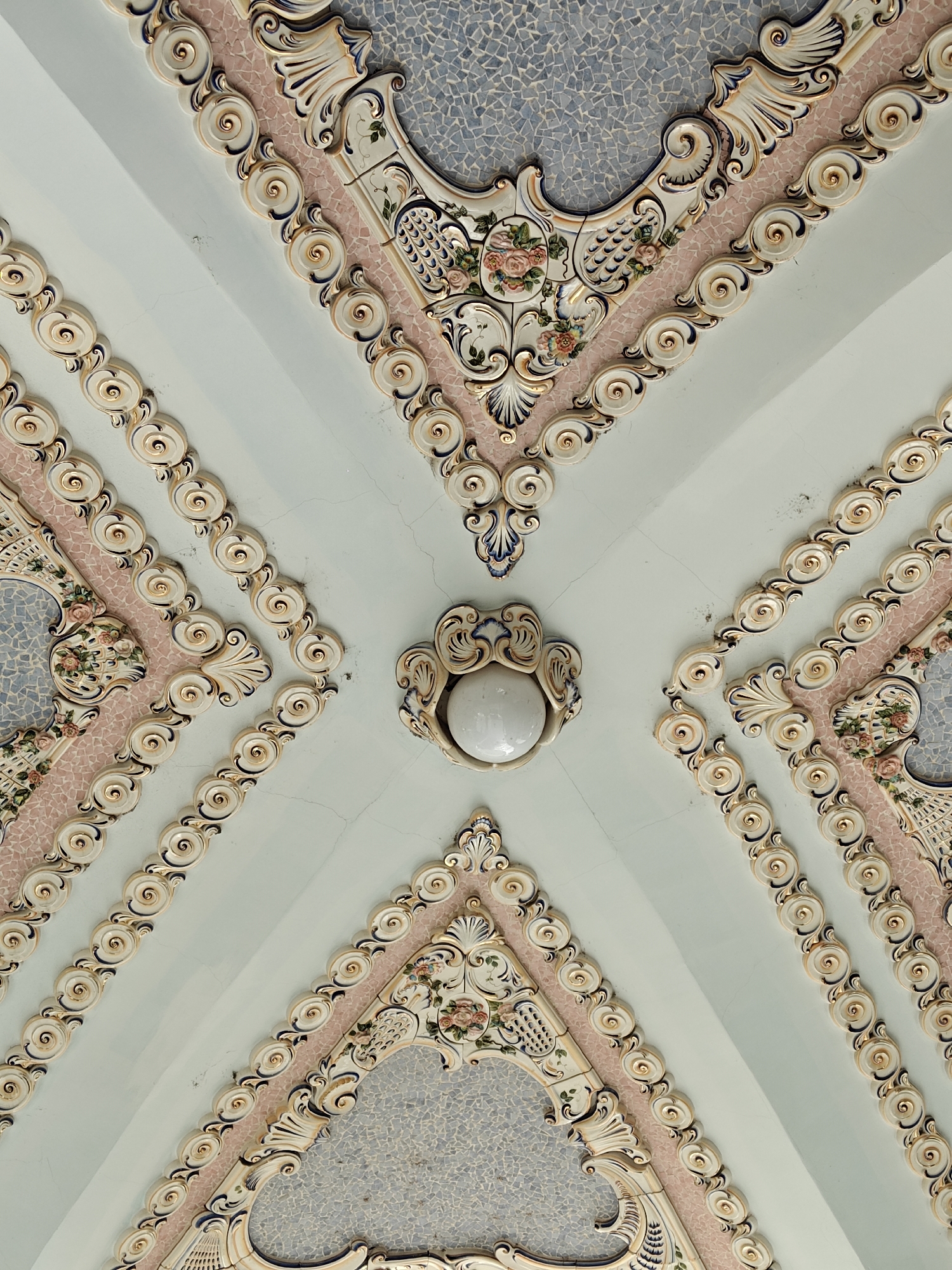 The image depicts a view of an ornate ceiling, likely indoors, located in the city of Batumi, Georgia. The ceiling is divided into four sections, each separated by a light blue field that converges at the center, where there is a stylized white lamp fixture. Each section features a decorative mosaic pattern in gray-blue and pink, surrounded by cream-colored ornamentation with floral motifs and scrolled edges in shades of white, pink, and light blue. There are decorative circular elements that run along the sides of each section. The ceiling has a vintage, possibly historical, aesthetic, with some visible cracks on the blue painted surfaces.