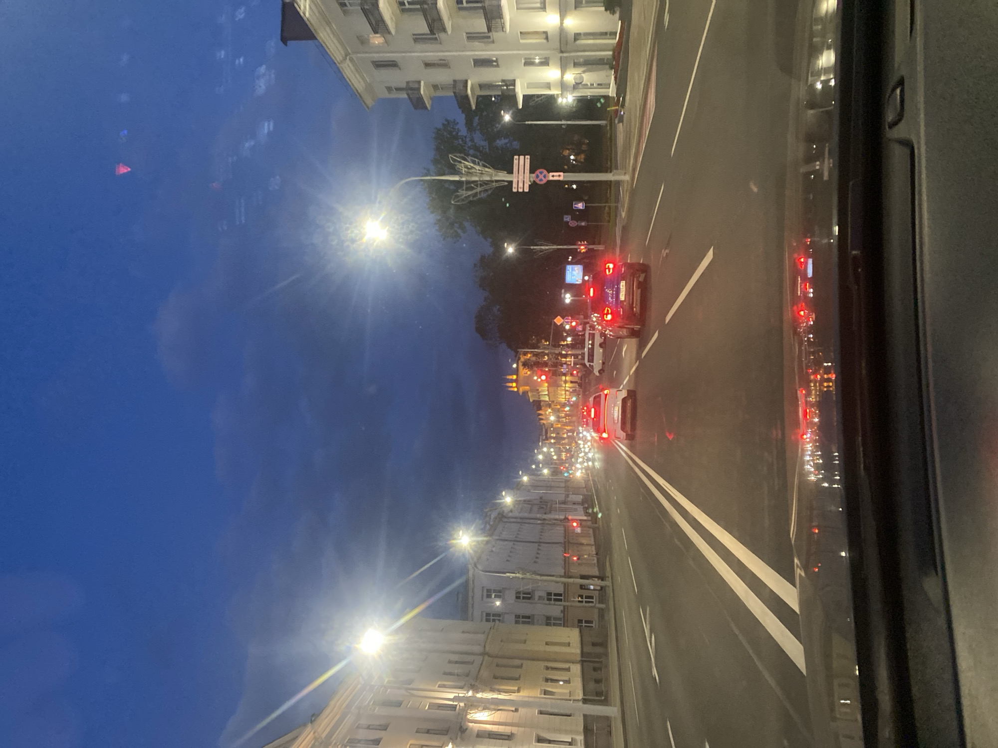 The image captures a street scene in Minsk, Belarus, at dusk. The perspective is from inside a vehicle, looking forward through the windshield. A two-lane road with white markings stretches ahead, flanked by multi-story buildings on both sides. Streetlights illuminate the scene, casting a glow on the road and buildings. Several cars are visible on the road, their headlights and taillights creating streaks of light. The sky is a deep blue with some clouds. In the distance, illuminated buildings and lights suggest a city center. Traffic lights are visible, some showing red. The overall impression is of a city at the transition from day to night.