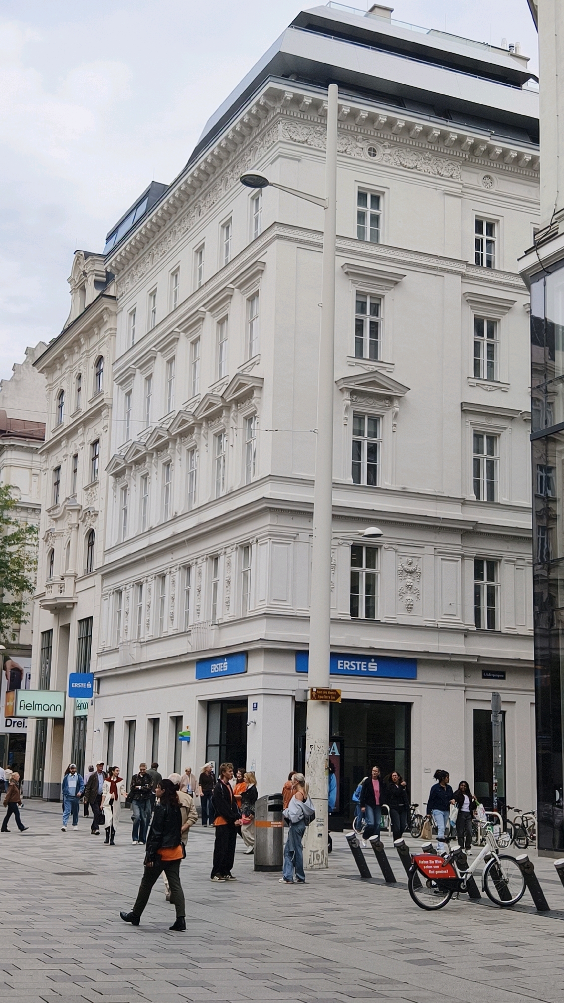 The image depicts a street scene in Vienna, Austria, likely a commercial area given the presence of shops and pedestrians. The architecture is classic European, characterized by an ornate white building with multiple stories, featuring numerous windows, decorative moldings, and a mansard roof. A modern addition is visible at the top of the building, contrasting with the historical design.
On the ground floor, signage for "ERSTE S" is prominent, indicating a bank or financial institution. Adjacent to this, another shop sign reads "fielmann" and "Drei", suggesting optician and telecommunications services respectively. The street is paved with grey rectangular tiles.
A group of people are walking and standing on the sidewalk. Some are dressed casually, while others appear to be shoppers or passersby. There are bicycles parked nearby, and some individuals are interacting with each other. The overall atmosphere appears to be a typical urban environment on a cloudy day. A lamppost stands in the foreground, an essential piece of street furniture.