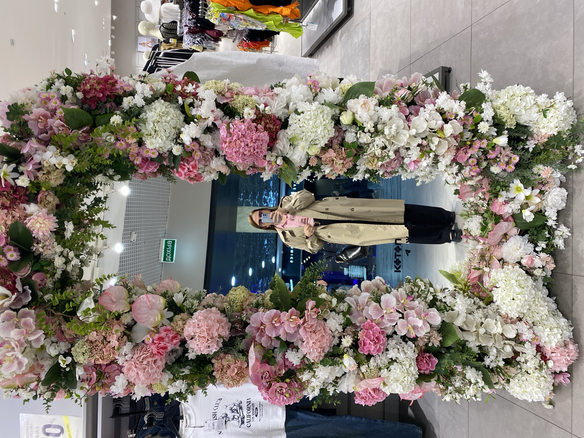 The image shows a full-length mirror surrounded by an elaborate floral arrangement of pink and white flowers. The mirror reflects a person wearing a tan trench coat, black pants, and a pink scarf. They are holding a smartphone, presumably taking a photograph of themselves.
The mirror is located inside a store. There are clothes racks visible behind the floral arrangement, and other mannequins and clothing items in the background, suggesting a retail environment. The store appears to be in Yekaterinburg, Russia, based on visible Cyrillic text ("ВЫХОД" meaning "Exit" and other Cyrillic script). The floor is tiled.