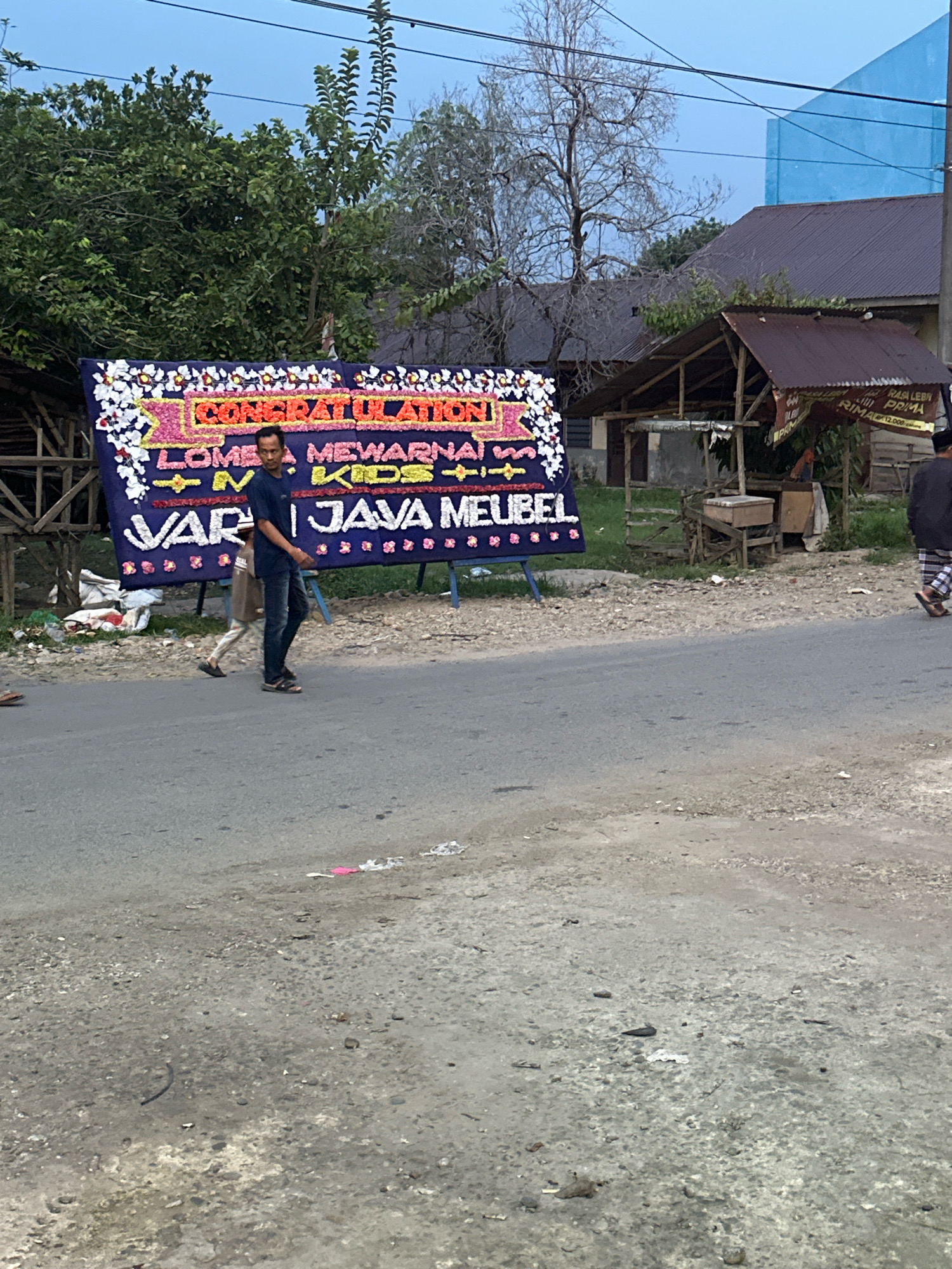In the city of Binjai, Indonesia, a large floral arrangement with the text "CONGRATULATION" and "JAVA MEUBEL" is displayed outdoors next to a road. A man in a dark blue t-shirt and jeans is standing next to the arrangement, looking towards the camera. Another person, partially visible and wearing lighter-colored pants, walks behind him. The floral arrangement is adorned with various flowers and has a dark blue banner. In the background, there are trees, a rustic wooden structure with a corrugated metal roof, and a building with a blue facade. The ground in the foreground is a mix of asphalt and gravel, with some debris scattered around. The sky is overcast.