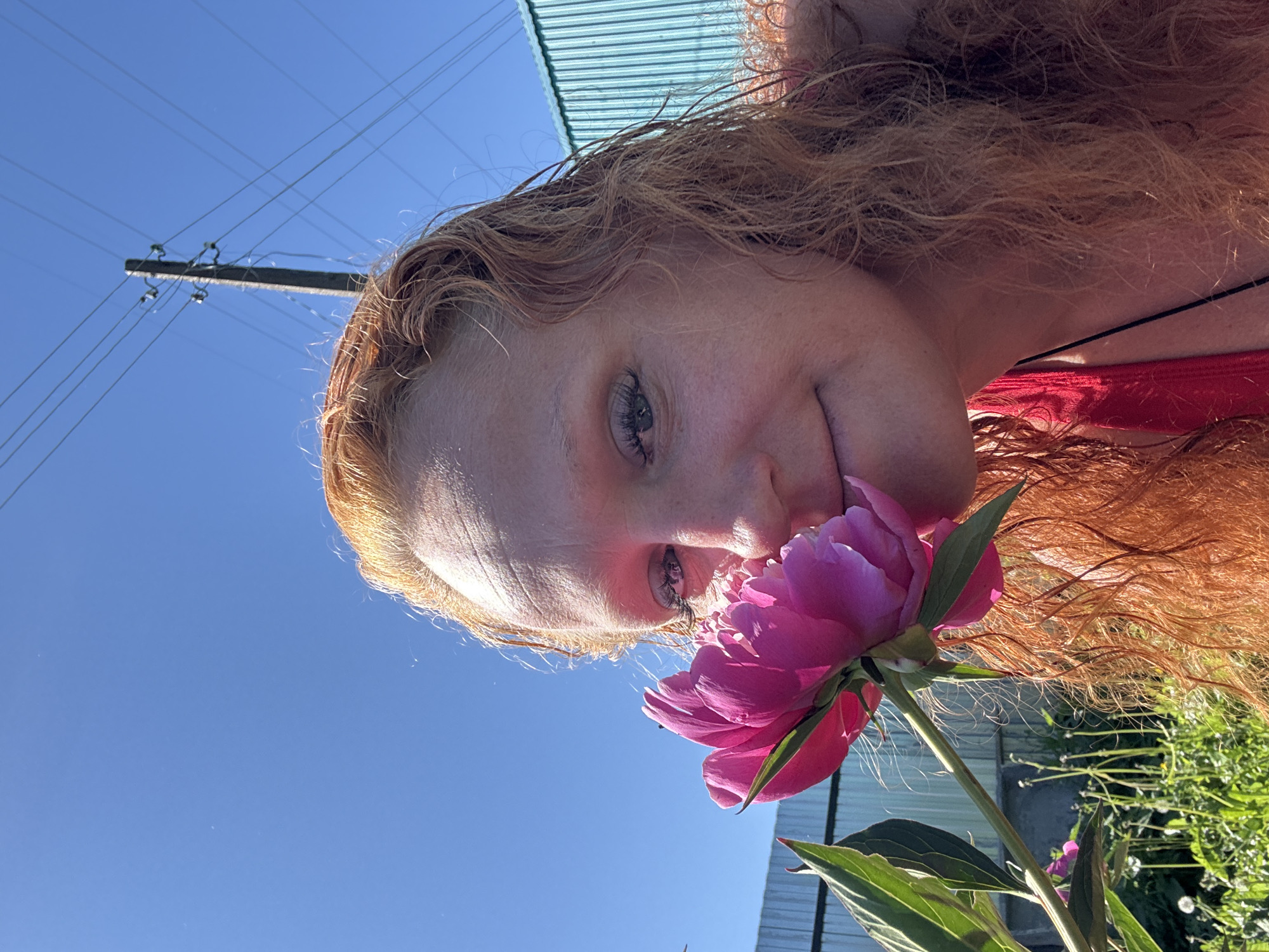 A woman with long, wavy, red hair is shown in close-up, smelling a bright pink peony flower. She is looking towards the camera with a slight smile. The background shows a bright blue sky with power lines and a utility pole. To the right is a light blue corrugated metal structure, possibly a building or fence. To the left, a green metal fence is visible, along with foliage, dandelions, and other plants in what appears to be a garden or yard. The scene is illuminated by bright sunlight. The location is likely an outdoor area within the city of Chaikovsky, Russia, suggested by the presence of typical urban infrastructure like power lines and utility poles.