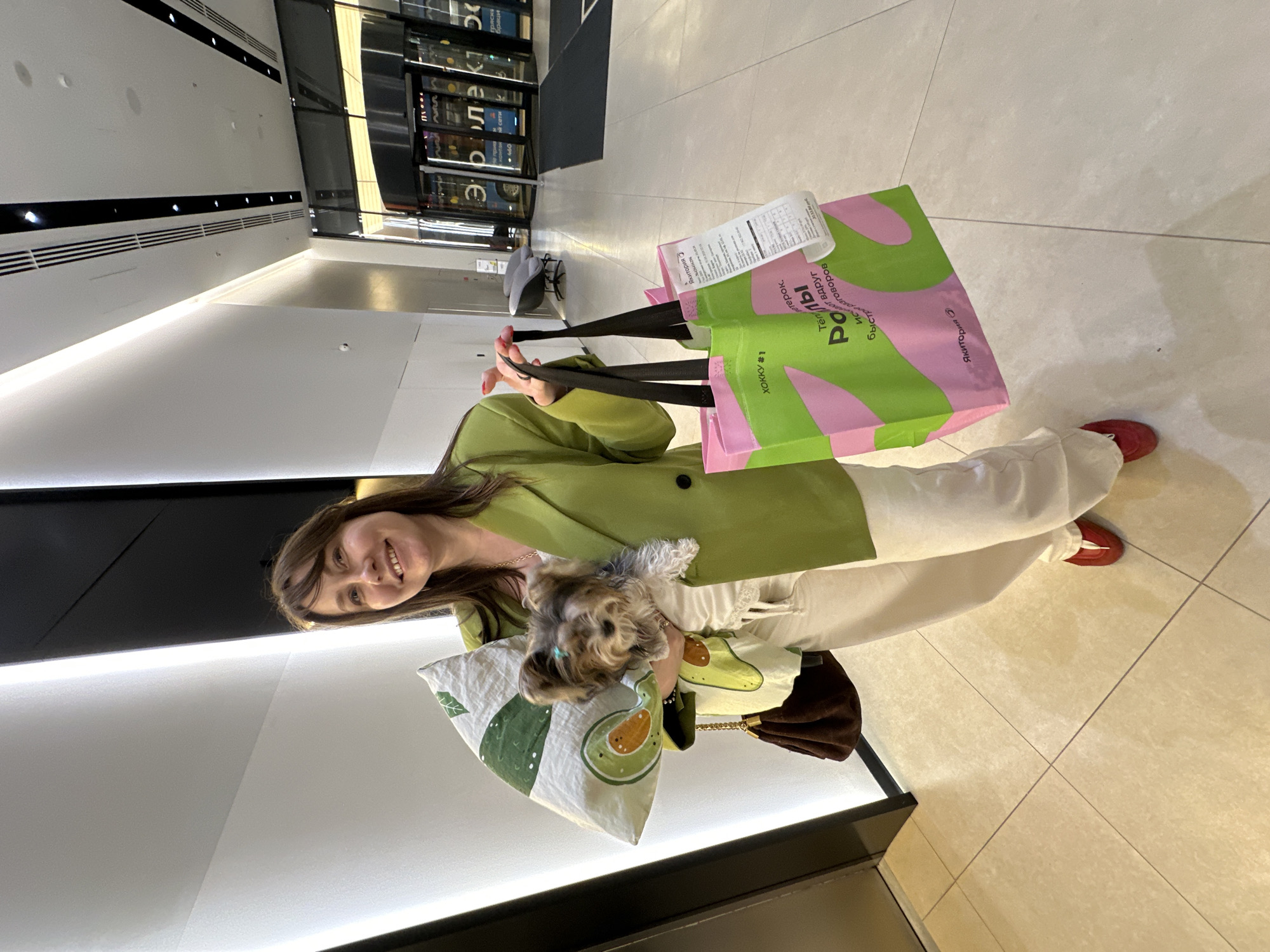 A woman in a light green blazer and white pants is holding shopping bags, a pillow with an avocado design, and a small dog in a matching avocado-themed garment. She is smiling at the camera. The shopping bag is from a store called "Teremok" and contains a receipt, indicating a purchase was made. The location appears to be a brightly lit indoor shopping mall, with tile flooring. The text on the shopping bag is in Russian. The presence of the "Yakitoriya" logo suggests a chain restaurant in Russia. The presence of Russian text and "Yakitoriya" suggest a location in Moscow, Russia.