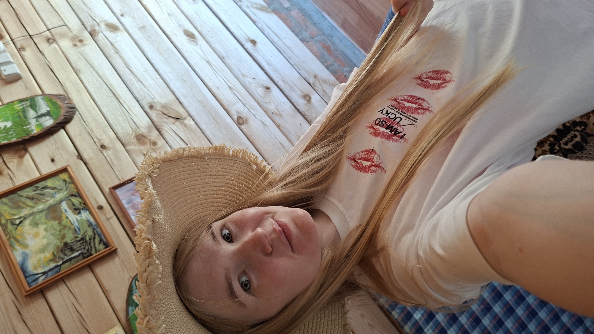 A medium shot of a young woman wearing a straw hat and a white T-shirt with red lipstick marks and the phrase "I AM SO LUCKY" lying on a blue and white checkered surface, possibly a blanket or cushion. The woman has long, light blonde hair and is looking directly at the camera with a slightly tilted head. The background includes a light wooden floor with several framed paintings or decorations along the left edge. The location is identified as Большой Букор, Russia.