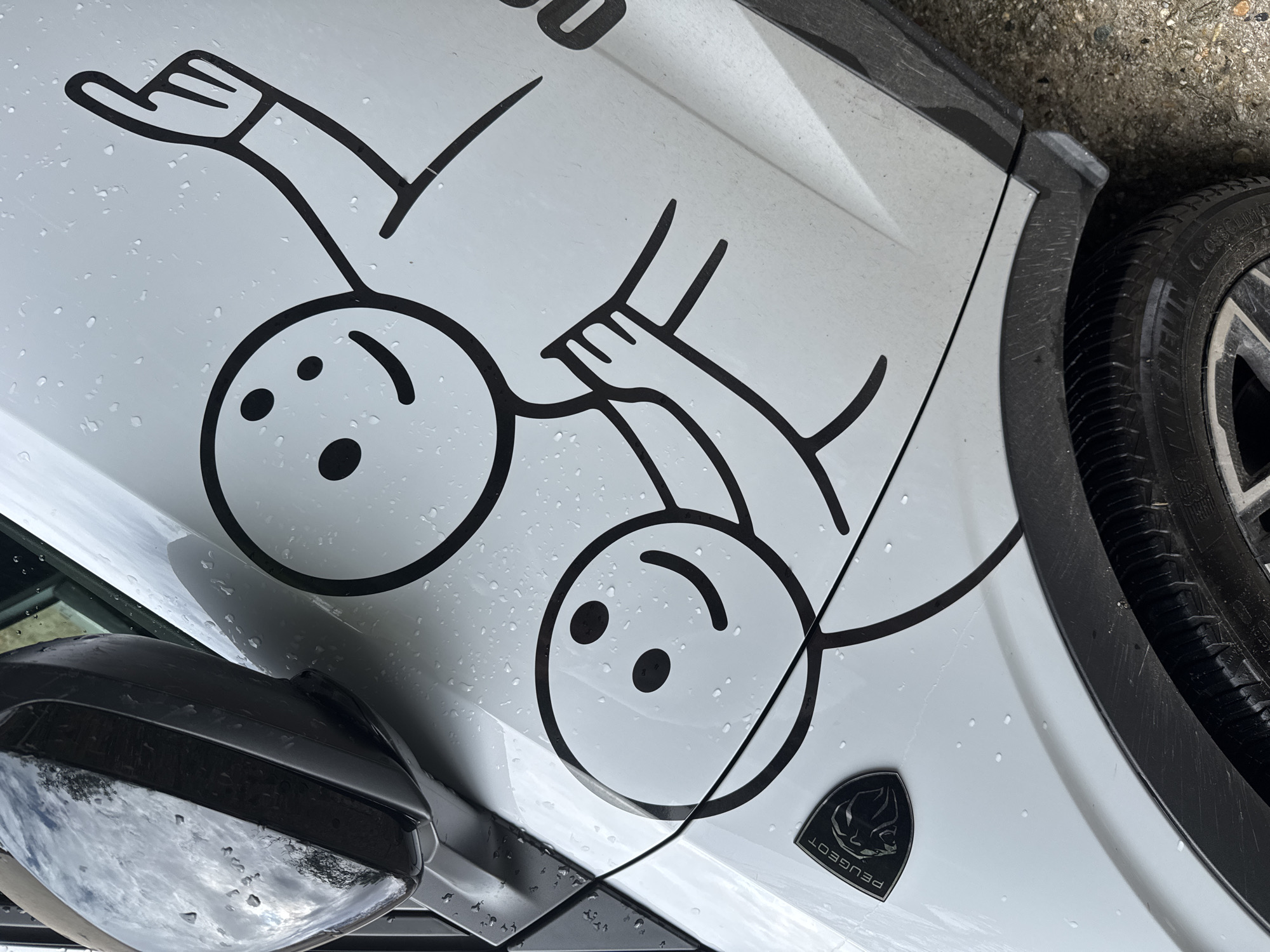An overhead, diagonal view captures the side of a white car, recently exposed to rain, evidenced by numerous water droplets on its surface. The primary focus is a large, black outline decal featuring two stylized, stick-figure-like characters on the car's body.
One character, positioned upright, has a round head with two small, black, oval eyes and an upward-curved line for a mouth, giving a smiling expression. Its left arm is raised, with the hand showing four fingers and a thumb in a "high-five" or waving gesture. The second character, placed slightly below and to the right, is depicted upside down relative to the first. It also has a round head with similar eyes and a curved mouth, appearing to smile. Its right arm is raised, with the hand mirroring the gesture of the first character, suggesting they are touching or interacting with hands. The figures are simplistic, with thick black outlines against the glossy white car surface.
Additional car details are visible: a black side mirror in the lower left, reflecting a cloudy sky and possibly trees. Towards the right, a portion of a car wheel is seen, featuring a dark rim and a tire with a distinct tread pattern. The tire sidewall faintly displays text, including "CROSSCLIMATE." Near the lower right of the decal, a black, shield-shaped emblem with the word "PEUGEOT" inscribed (upside down in the image's orientation) is present, featuring the brand's lion logo. The ground next to the car tire appears to be a concrete or paved surface. Faint, dark numbers like "00" are partially visible above the left character's head, possibly part of another decal or text on the car. The lighting suggests it is daytime, likely overcast due to the rain.