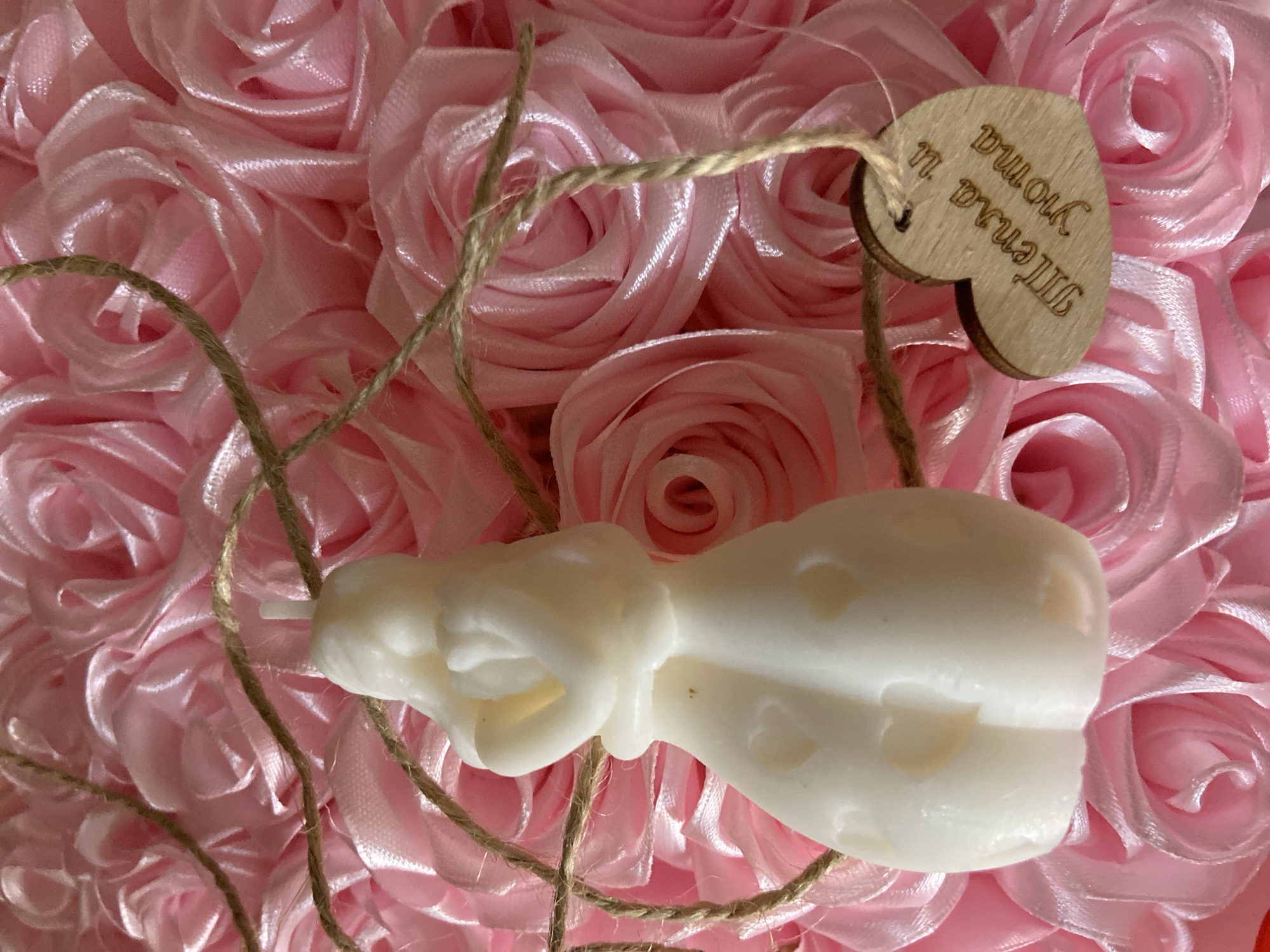 The image shows a close-up of a white candle shaped like two figures embracing, standing on a bed of pink satin roses. A rustic twine cord is draped around the candle and a small wooden heart charm is attached to it. The wooden heart is engraved with text that appears to be in Cyrillic script, reading "Тепла и уюта". This translates to "Warmth and comfort". The background is entirely filled with the pink roses, which are tightly packed and create a textured, floral backdrop. The lighting is soft and diffused, highlighting the details of the candle and the texture of the roses. There are no people or animals visible in the image. The scene evokes a sense of romance, warmth, and domesticity, likely intended as a gift or decoration. It is not possible to determine the location in the city of Япрыково, Russia, from the image alone.