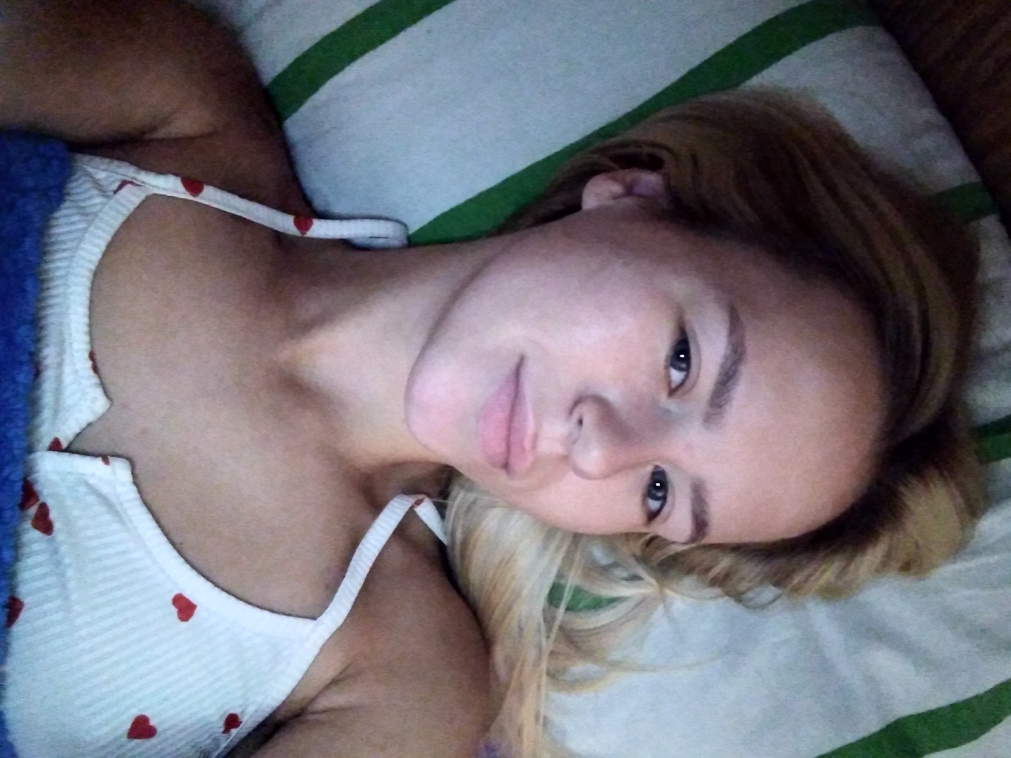 A young woman with blonde hair is lying in bed. She is looking at the camera with a neutral expression. She is wearing a white tank top with red hearts printed on it. The bed is covered with a striped blanket. The scene appears to be indoors. No specific location in Radisne, Ukraine can be identified.