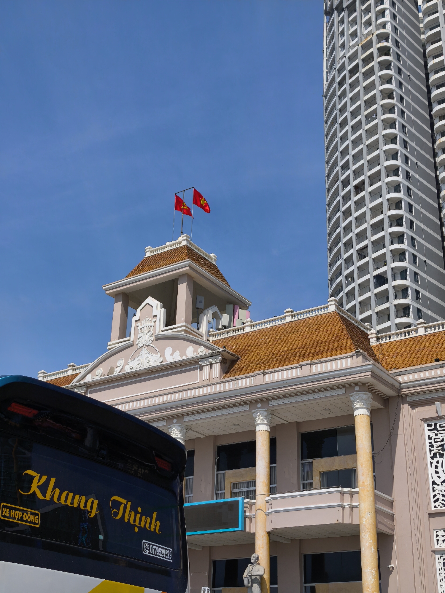 The image captures a sunny daytime scene in Nha Trang Ward, Vietnam. In the foreground, the rear of a dark blue bus is visible, displaying the name "Khang Thịnh" in yellow cursive lettering. To the left, a small yellow rectangle with the text "XE HỢP ĐỒNG" is present. On the right side of the bus's rear window, a white rectangle shows a phone number.
Behind the bus, a light pink building with a classic architectural style is prominent. It features an orange-tiled roof, decorative white trim, and large columns. Two red flags with yellow stars, the national flags of Vietnam, fly from a flagpole atop the building's central tower. Adjacent to this building, a modern, tall, grey skyscraper with a repeating pattern of balconies rises into the clear blue sky. A statue of a person in a suit is visible at the base of the pink building. The overall environment suggests an urban setting with a mix of historical and contemporary architecture.
