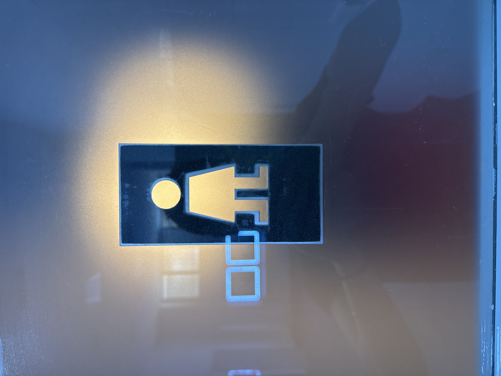 An illuminated, dark blue and gold-colored restroom sign featuring a female silhouette is affixed to a neutral-toned, reflective surface. The sign is rectangular with rounded corners and is positioned slightly off-center to the right. To the left of the female silhouette are two glowing blue, square-like shapes. The reflective surface shows a distorted reflection of overhead lights, creating a halo effect around the sign. The bottom portion of the image reveals a darker, red-tinted reflection, possibly of the photographer or a person nearby, along with a bright blue horizontal line at the very bottom, suggesting a floor or a lower surface. The scene is indoors, likely on a door or wall in a public building. There is no discernible information in the image to indicate the specific location within the city of Murcia, Spain.