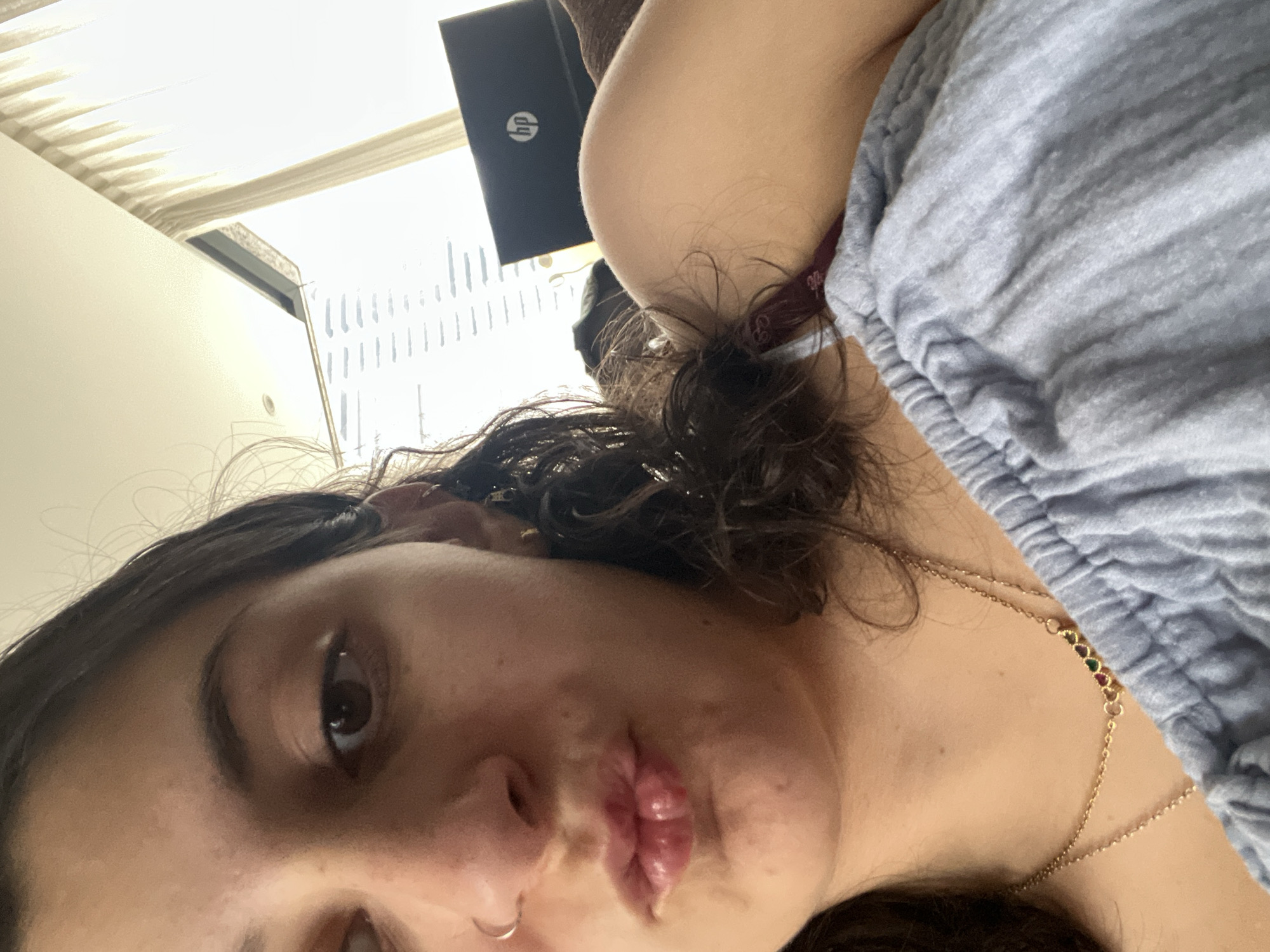 A woman lies on a bed, facing the camera with pursed lips. She has dark curly hair, fair skin, and wears a nose ring and a gold necklace with colorful stones. She is partially covered by a light gray blanket or garment. Behind her, a black HP laptop is visible near a window with vertical blinds. Based on the window blinds, it appears the location is within Mexico City, Mexico.