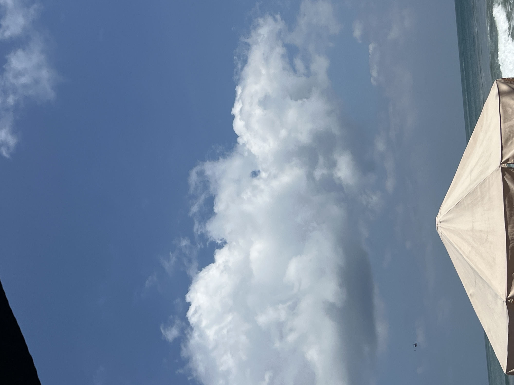 A wide-angle, upward-looking shot shows a bright blue sky with scattered white clouds. The lower part of the frame is occupied by the cream-colored fabric dome of a large beach umbrella, with its edge visible at the bottom. To the left of the umbrella, the deep blue ocean is visible with waves breaking on the shore. In the distance, a small, dark helicopter flies horizontally across the sky. The scene appears to be a beach in Kuta, Indonesia.