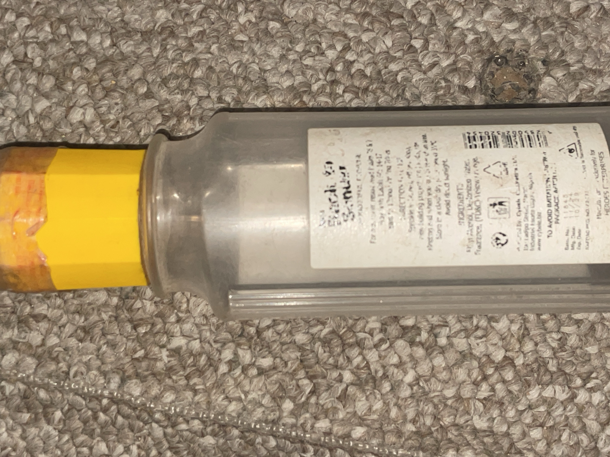 The image shows a close-up of a plastic bottle with a yellow cap, resting on a textured carpet. The bottle has a label with text, some of which is legible. The text on the label appears to be product information, including ingredients, directions for use, and manufacturer details. The bottom of the label indicates it is manufactured by "Cybelk Cosmetics Ltd." in Jalingo, Nigeria. The surrounding environment is a carpeted surface with visible texture and some debris.