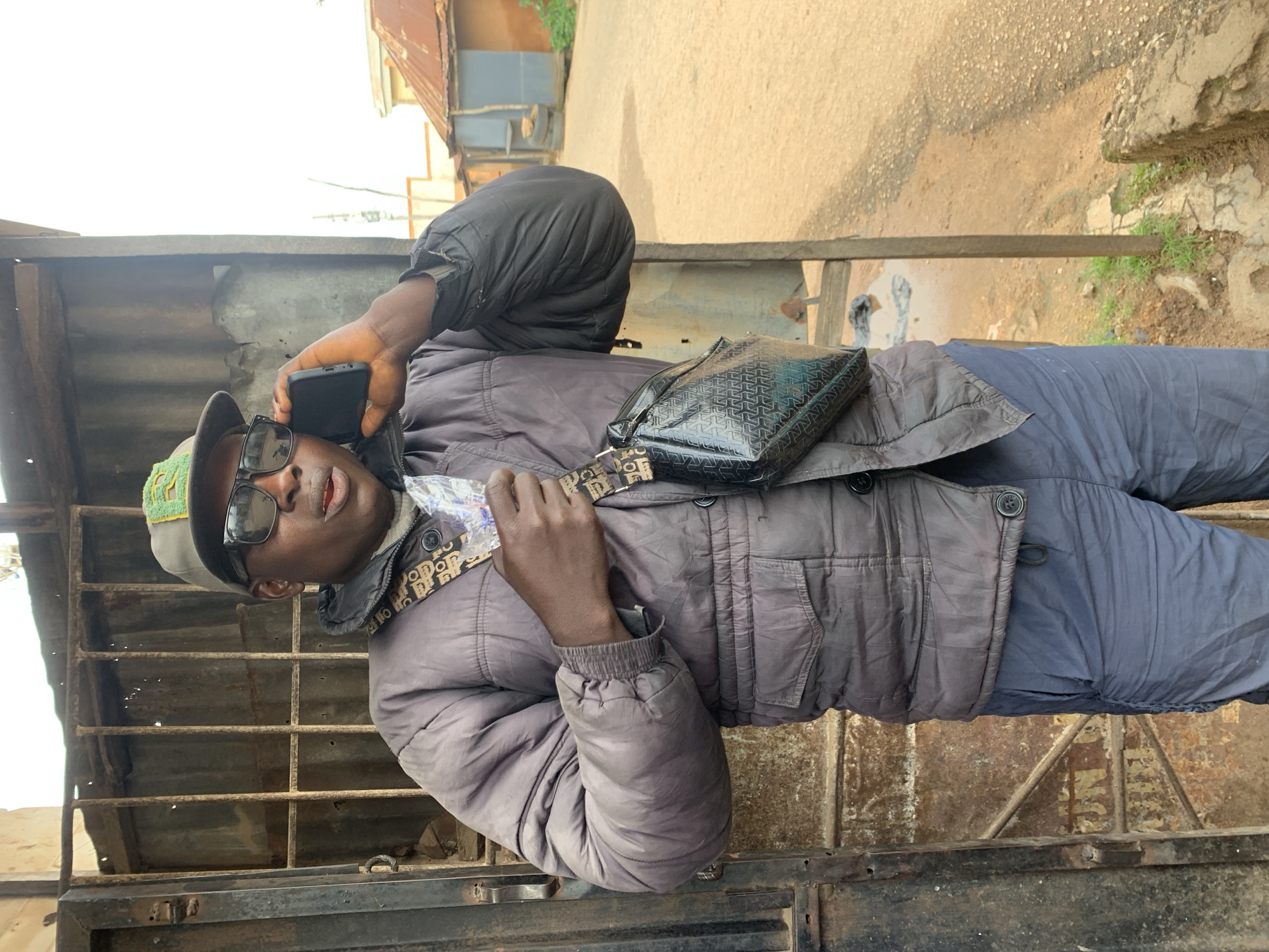 A man stands outdoors in an urban environment, talking on a mobile phone. He is wearing a grey baseball cap with a green emblem, dark sunglasses, a grey puffer jacket over a lighter grey garment, and dark blue trousers. A black crossbody bag with a repeating pattern is slung across his chest. He is holding a small item, possibly a card or ticket, in his right hand. The background includes a rusty metal gate or fence, a corrugated metal structure, and a dirt road with buildings and vegetation. The overall scene suggests a casual setting in a developing area.