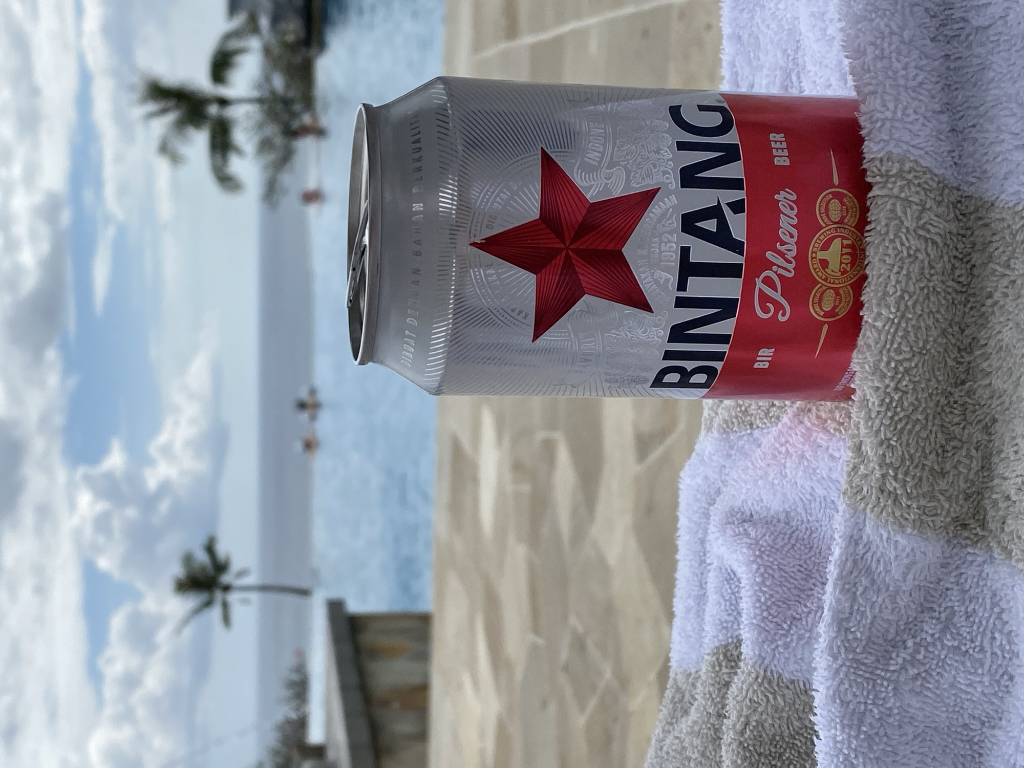 A Bintang beer can sits on a striped towel next to an infinity pool. In the background, palm trees and the ocean are visible, with a cloudy sky overhead. The can has a red star logo and text indicating "BIR Pilsener BEER". The poolside area has stone paving. Several people are faintly visible in the pool in the distance. The scene appears to be a resort in Kuta Selatan, Indonesia, possibly a beachfront property given the ocean view.