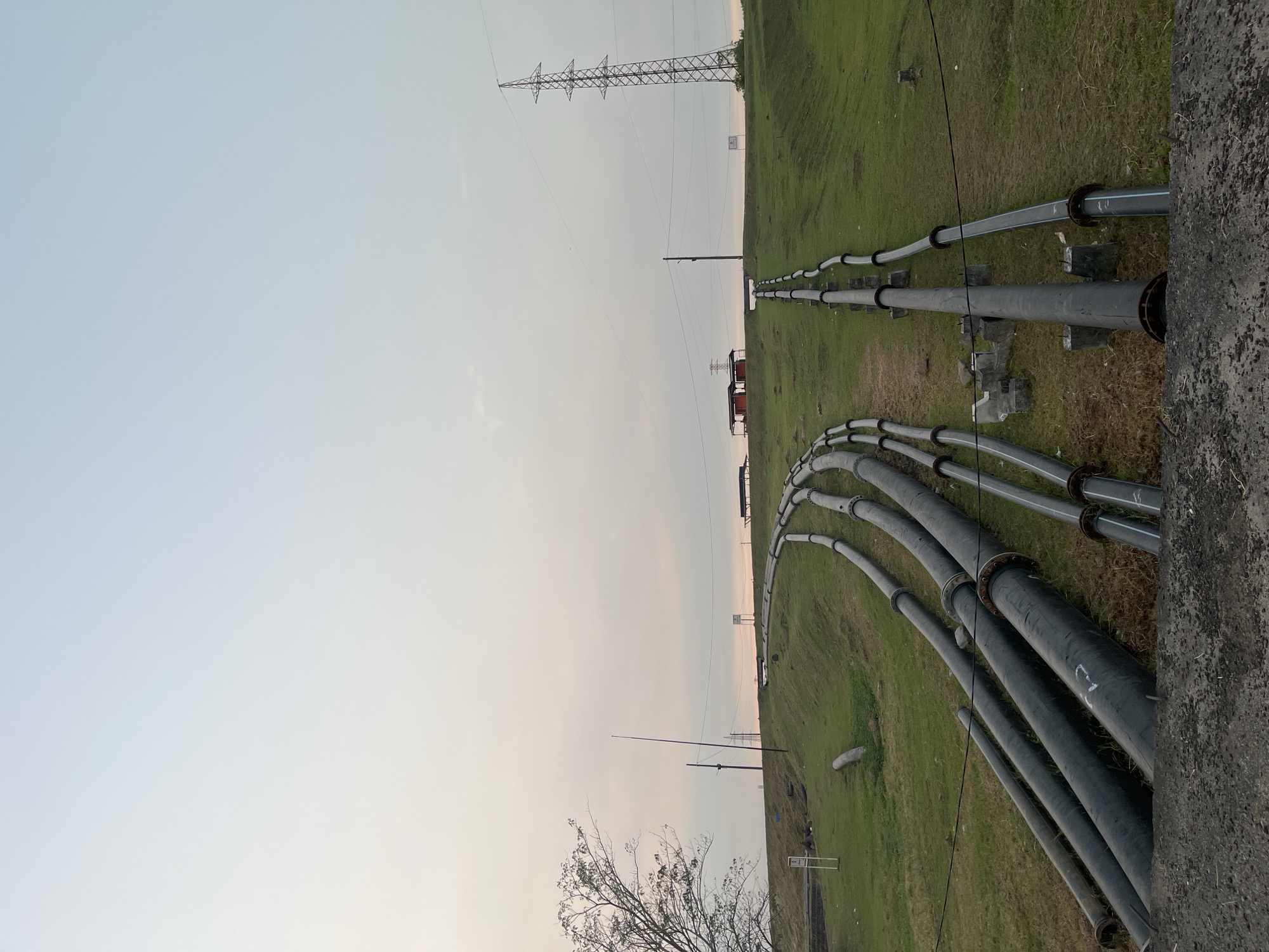The image depicts an industrial landscape in Besuki, Indonesia, characterized by a series of large, dark pipes laid across a grassy embankment. The pipes, some appearing to be made of metal and others of a synthetic material, are arranged in parallel lines and curve upwards along the slope. They are supported by concrete structures and metal stands, indicating their function in transporting fluids, likely for industrial or water management purposes.
In the background, a tall, lattice-like electrical pylon dominates the skyline, suggesting the presence of power infrastructure. Several small, red-roofed buildings are visible near the crest of the embankment, possibly associated with the industrial operation. A lone tree with sparse foliage stands on the left side of the frame, while the sky is overcast with subtle hues of pink and orange, suggesting it might be dawn or dusk. The foreground is occupied by a rough, concrete surface, hinting at a man-made structure or pathway. No people are visible in the scene. The overall impression is one of functional infrastructure within a natural, albeit somewhat barren, environment.