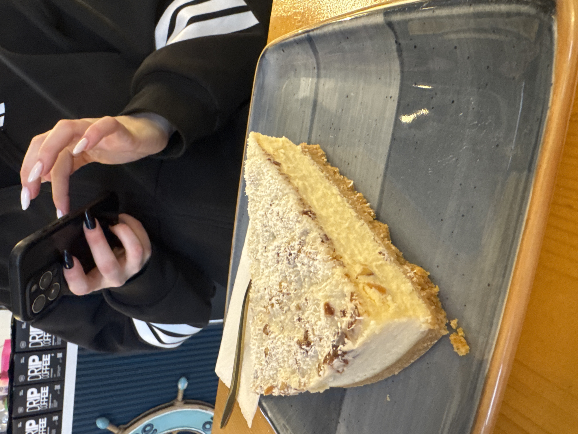A person wearing a black Adidas sweatshirt is holding a cell phone. A slice of cake is on a blue plate with a wooden edge. A fork is next to the cake. Boxes labeled "DRIP COFFEE" and a nautical-themed decoration are visible.