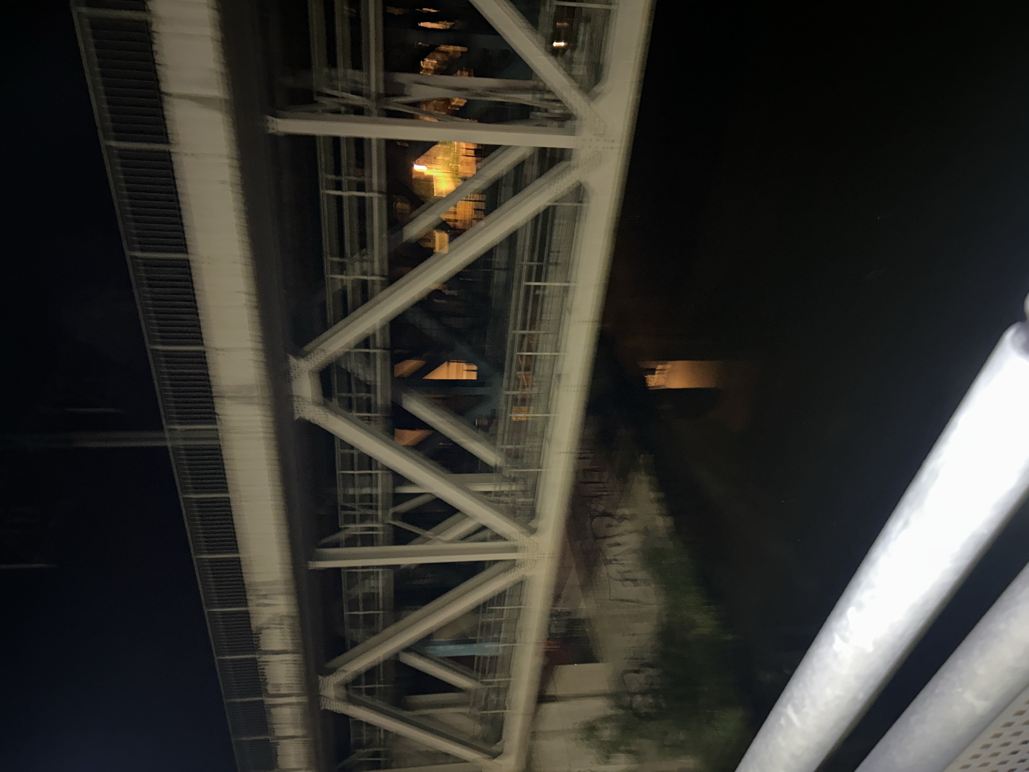 The image depicts a nighttime view of a bridge over a body of water, likely in Pilsen, Czechia. The bridge features a complex lattice structure made of white metal beams. The water below reflects the lights and structure of the bridge. The foreground shows part of a railing or walkway, also appearing to be made of metal. The overall atmosphere is dark and industrial, with the only significant illumination coming from the bridge's lights and perhaps some distant urban lighting. There are no people visible in the image.