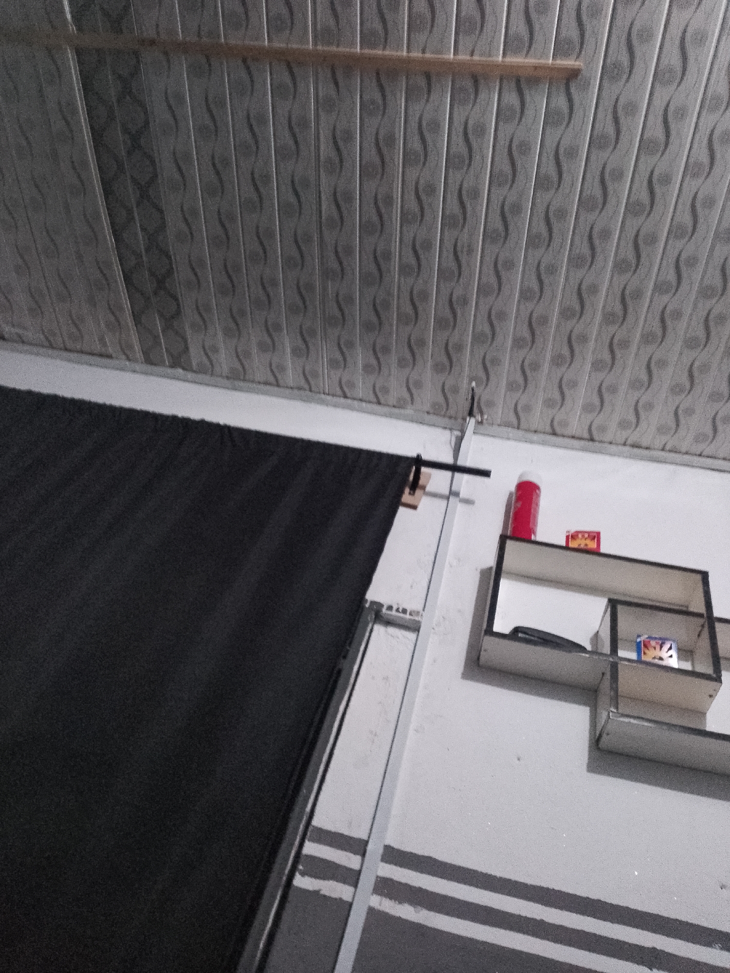 An indoor, low-angle shot shows a dark-colored curtain or fabric hanging on the left side of the image. Above the curtain, a white ceiling with a textured, wavy pattern is visible, interrupted by a thin, light-colored wooden beam running horizontally across the top. On the right side of the image, a white wall features a small, modern shelf unit with two levels. The top shelf holds a red cylindrical object and a small yellow and red box. The bottom shelf contains a dark object and another small box with colorful graphics. Below the shelf, the wall has three horizontal dark gray stripes. A metal support rod extends from the ceiling down the wall near the curtain. The overall scene appears to be in a room, possibly a storage area or a living space.