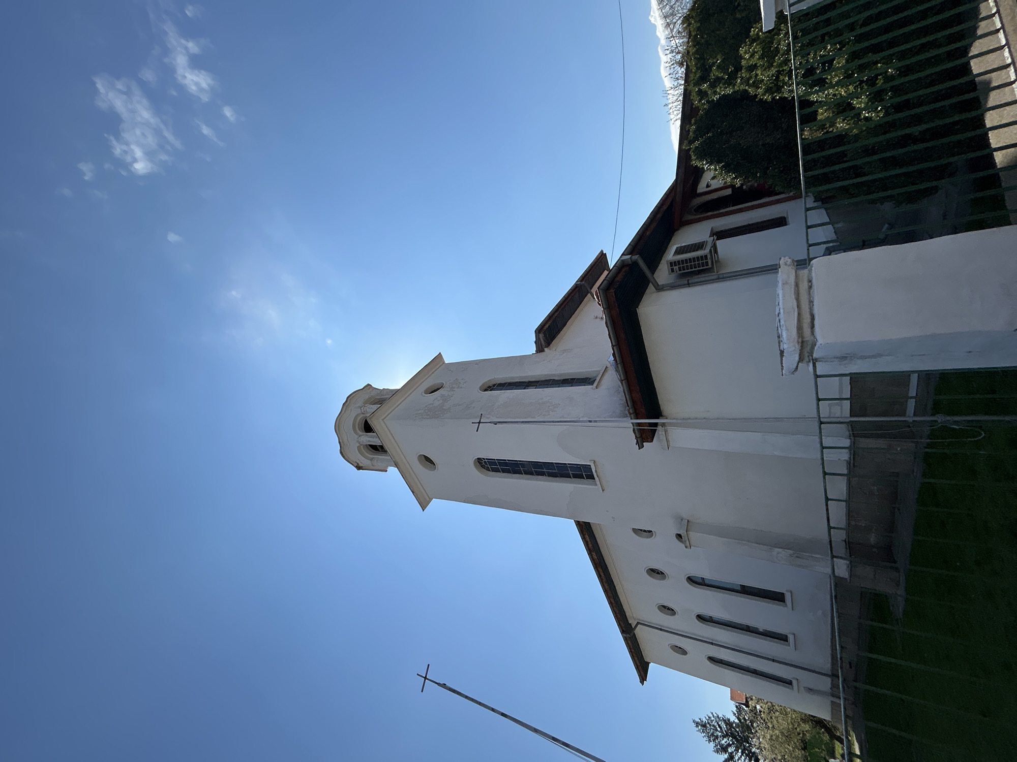 A low-angle, outdoor shot shows a white building with a tall steeple against a blue sky with wispy clouds. The building has multiple windows, a dark roofline, and an air conditioning unit on the side. A cross is mounted on a pole in front of the building. A fence and some greenery are visible on the right side of the frame. The building is the Catholic Cathedral of St. Louis in Plovdiv, Bulgaria.