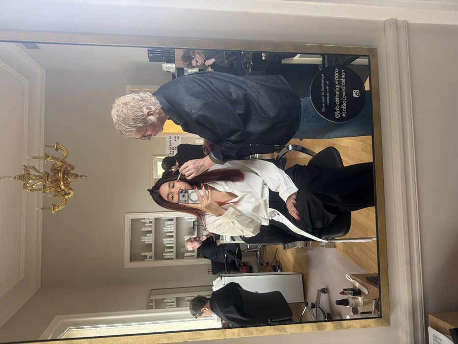 The image captures a detailed mirror reflection of a beauty salon interior in Paris, France, identified as a La Biosthetique establishment by a circular sticker on the mirror. A young woman with long, reddish-brown hair and red fingernails, dressed in a white collared shirt and black wide-leg trousers, is seated and actively capturing a mirror selfie with her smartphone. To her immediate right, an older individual with short, light-colored hair and glasses, wearing a denim jacket and jeans, meticulously applies makeup to the woman's face using a small brush.
The reflected environment reveals a well-lit room with light-colored walls, a white ceiling adorned with decorative molding, and an ornate gold chandelier suspended centrally. In the background reflections, shelves displaying numerous identical white cosmetic product bottles are visible. Additional individuals are reflected: a person in a black smock seated to the left, and a bearded man seated further back. On a counter in the lower-left corner of the reflection, various makeup products including lipstick and brushes are arranged. The circular black sticker on the mirror displays the text "Share your La Biosthetique moments with us! @labiosthetiqueparis #LaBioloversFashion" and an Instagram logo.