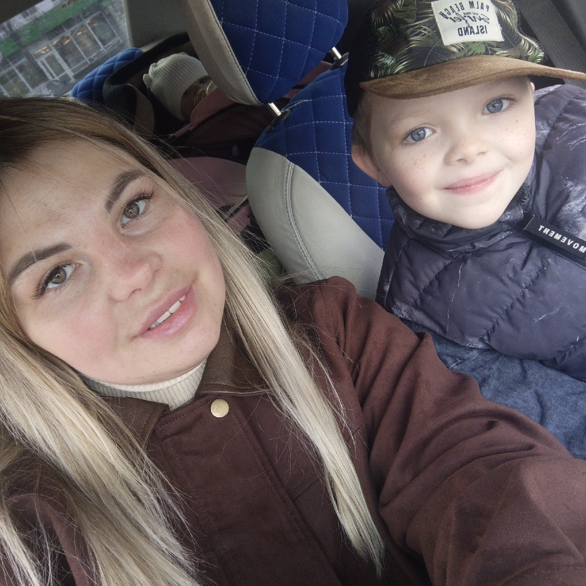 A selfie taken inside a car features a woman, a young boy, and an infant, capturing a casual travel moment.
On the left side of the frame, a woman with long blonde hair and light brown eyes smiles faintly towards the camera. She wears a brown jacket with a leather collar over a light-colored turtleneck sweater.
To her right, a young boy with bright blue eyes and freckles smiles broadly. He is wearing a dark blue or black quilted puffy jacket. On his head, he sports a camouflage-patterned baseball cap with a beige brim. A white label on the cap shows mirrored text that reads "ISLAND SURFERS BEACH PALM". A tag on his jacket also displays the mirrored word "MOVEMENT".
In the background, behind the boy and partially obscured, an infant or baby is visible in a car seat, wearing a white knitted hat and what appears to be a pacifier.
The car interior features blue quilted headrests and light-colored trim on the seats. Through the car window in the upper left background, an outdoor scene is reflected, showing parts of buildings or structures, with some mirrored text that is difficult to decipher. The lighting suggests it is daytime.