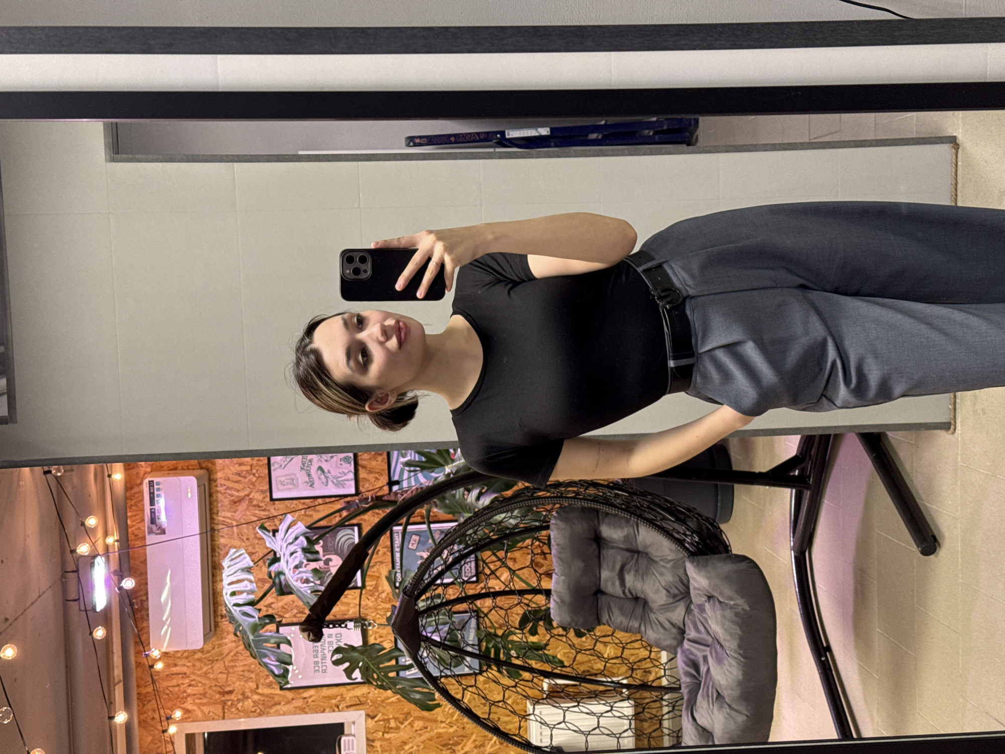 A young woman is captured taking a mirror selfie in an indoor setting. The image is oriented vertically with the woman on the left, holding a black smartphone with her right hand to photograph her reflection. She is wearing a black short-sleeved top, dark grey or blue trousers, and a black belt. A ring is visible on the fourth finger of her left hand. Her expression is neutral to slightly smiling, and her dark hair is pulled back, with some strands framing her face.
The scene reflected in the mirror shows a wall with light-colored, possibly tiled, surfaces and a dark, narrow horizontal frame or fixture above. A blue object, possibly a folded ladder or a shelf, is also partially visible in the upper reflected area.
Behind the mirror, the actual background reveals a distinctly decorated room. The wall is covered in light-colored wooden panels. Various decorative elements are arranged against this wall, including a large Monstera plant. To the left, a white air conditioning unit is mounted, displaying what appears to be a temperature setting, possibly "20.1°C". Below and around the air conditioner, strings of warm-toned bulb lights are draped, creating an inviting ambiance. Several framed pictures or posters are also visible on the wooden wall, though any text on them, such as "ED BEC", is mostly illegible. In the lower-right foreground, a black metal hanging egg chair with a plush grey cushion provides a comfortable seating option, partly obscured by the woman's body.
The overall atmosphere suggests a modern, possibly commercial space like a cafe or a plant shop, characterized by its warm lighting and contemporary decor.