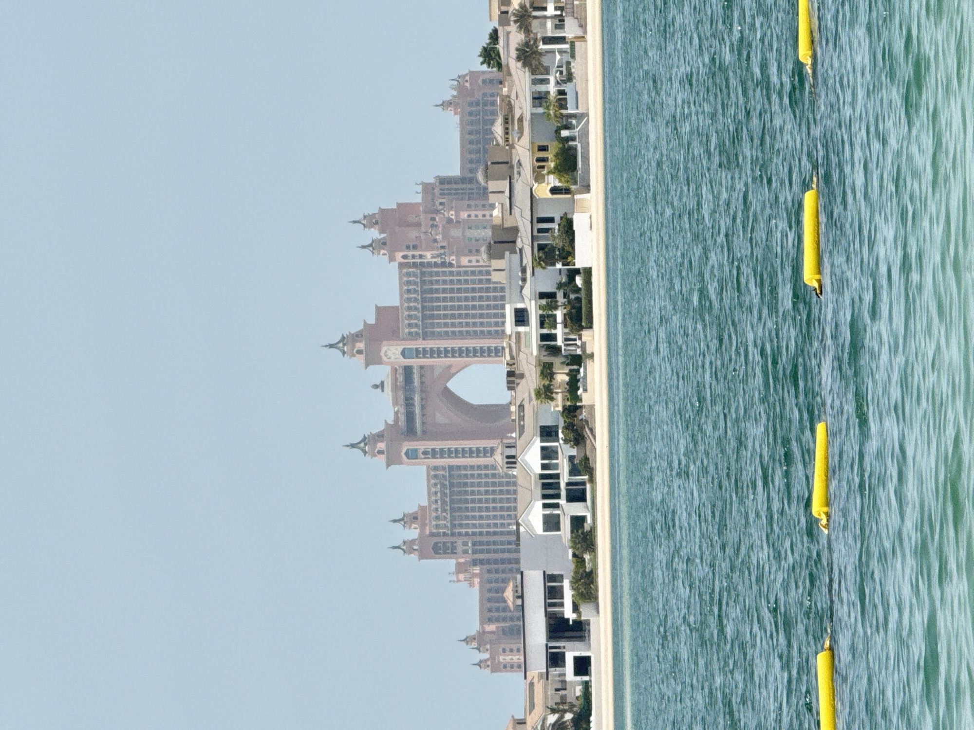 The image shows a view of the Atlantis, The Palm hotel and resort in Dubai, United Arab Emirates, from across a body of water. The resort is a large, pink-colored complex with distinctive architecture, including two towers connected by a central arch. In the foreground, several yellow buoys float on the surface of the turquoise water, likely marking a swimming area or boundary. Between the water and the resort, a strip of sandy beach is visible, dotted with palm trees and several modern, white villas with flat roofs and large windows. The sky is a hazy, light blue. The overall scene depicts a tropical resort environment with a clear view of a prominent Dubai landmark.