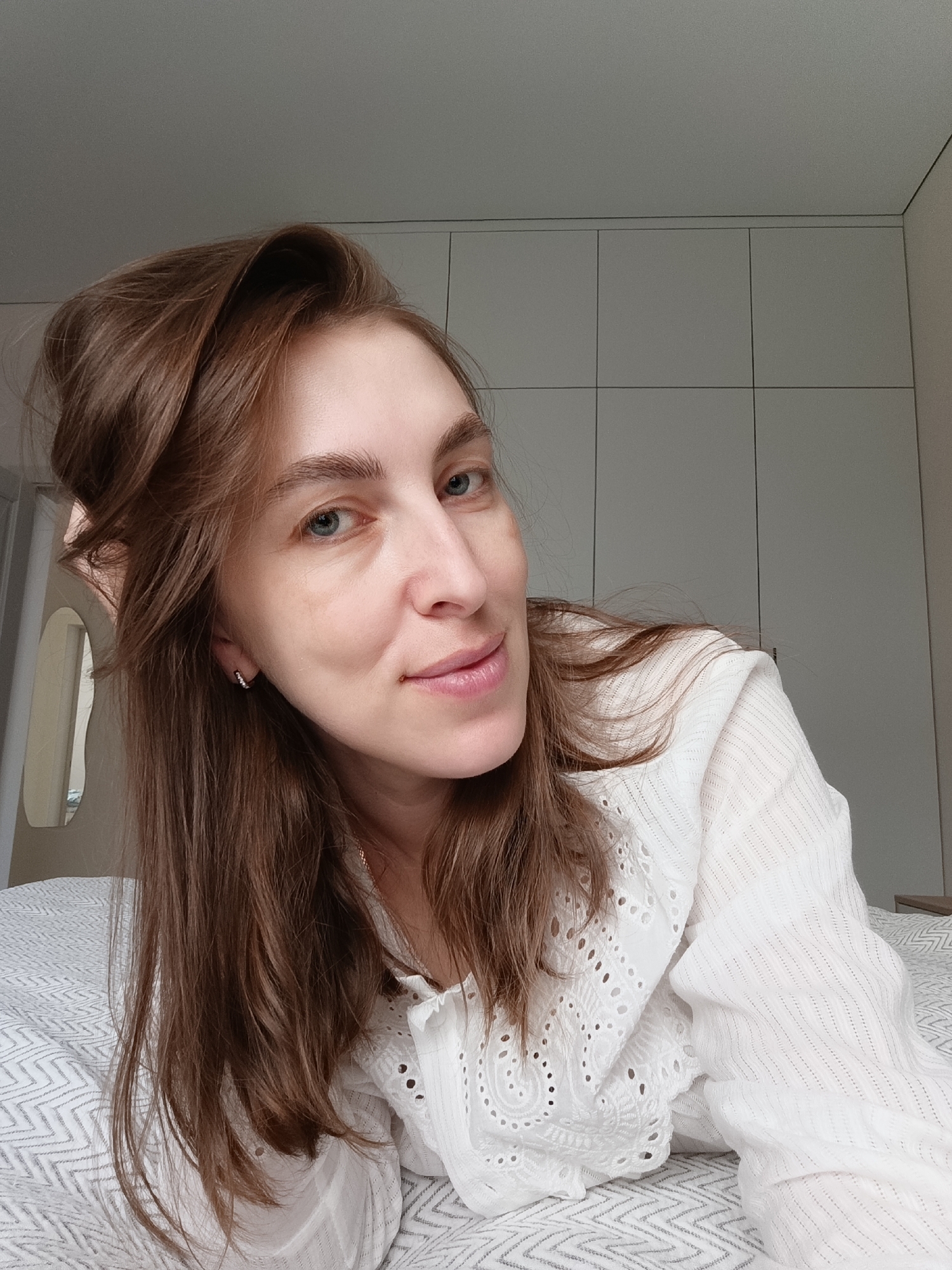 A woman with light skin and long, light-brown hair is lying on a bed covered with a white and grey patterned blanket. She is wearing a white blouse with embroidered eyelet details. Her hair is partially pulled back with her hand. The background consists of a light-grey wardrobe with a grid pattern of doors and a white door frame with a mirror. The location cannot be determined as Chaikovsky, Russia, based on the image alone.