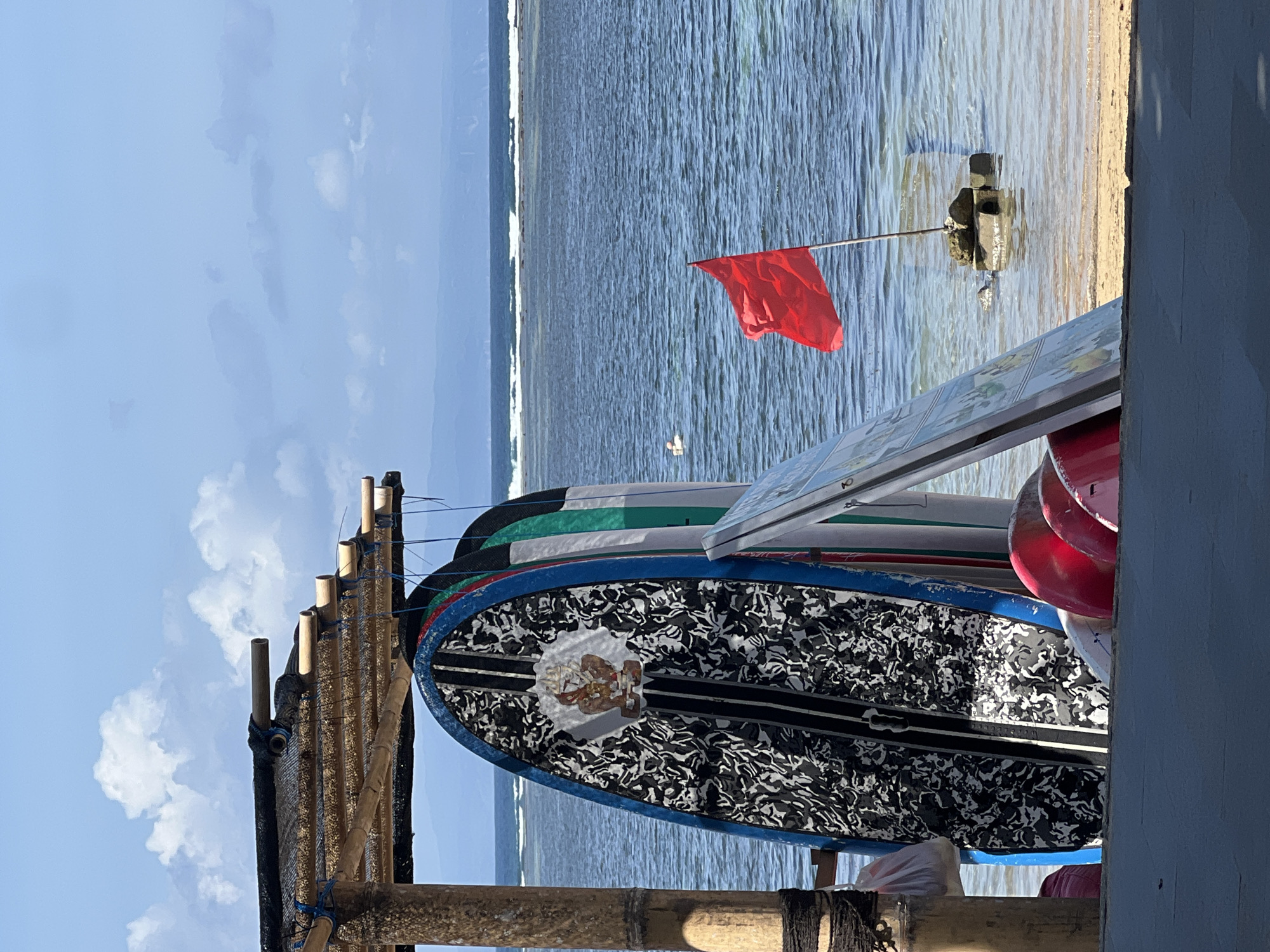 This image captures a scene at a beach, likely in the Denpasar area of Indonesia, given the presence of surfboards and the tropical atmosphere. A bamboo structure with a thatched roof provides shade. Several surfboards, including one with a black and white patterned design and a central emblem, are leaning against the structure. Some red surfboards are also visible at the bottom of the frame.
In the water, a red flag is planted on a submerged structure, signaling potentially hazardous conditions. The ocean stretches to the horizon, with gentle waves breaking. The sky is a clear blue, dotted with white, fluffy clouds. In the distance, a faint outline of land and mountains can be seen. A single person is visible in the water further out, appearing to be swimming or paddleboarding. The sand and shallow water are visible in the foreground.