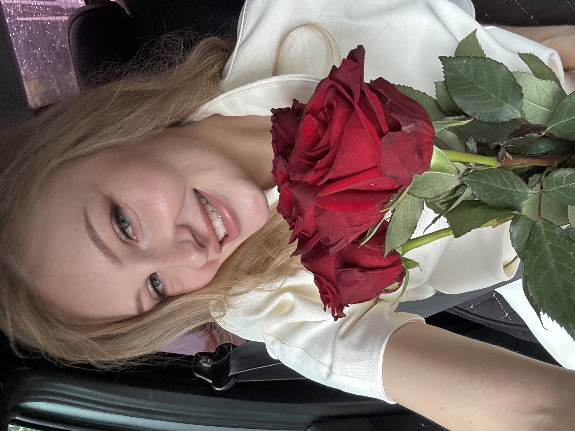 A young woman with blonde hair and fair skin is holding a bouquet of red roses. She is wearing a white jacket and is seated in a car. The background suggests an urban environment, possibly in the city of Perm, Russia, indicated by the visible car interior and a window with a rainy, blurred cityscape. Her expression is cheerful, and she is looking towards the camera.