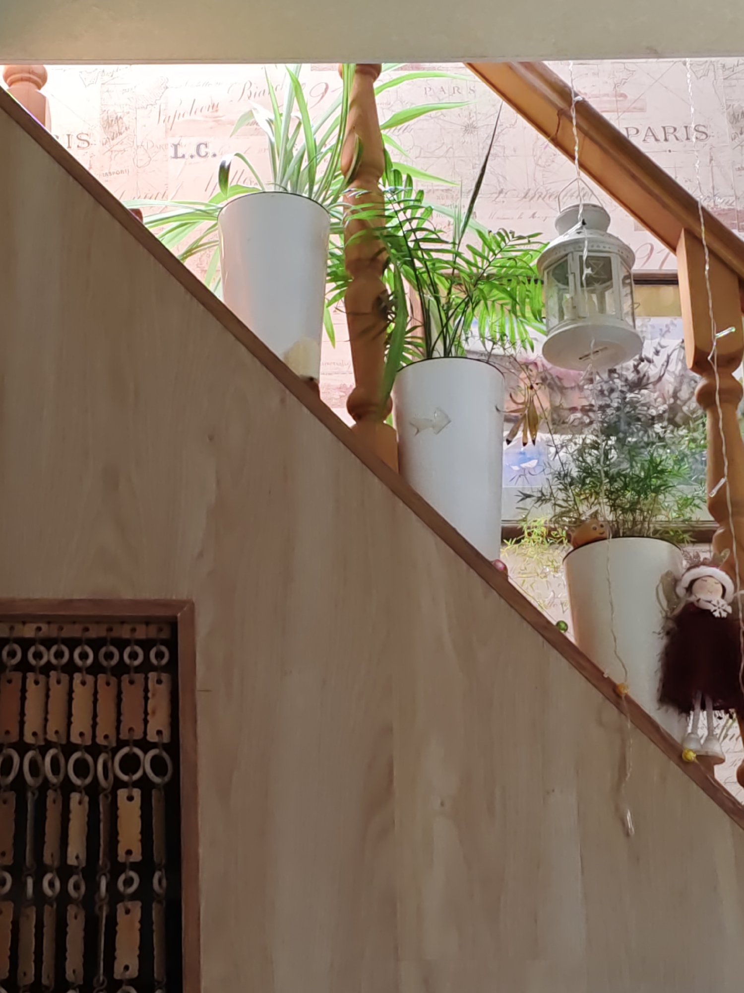 The image captures an indoor scene, likely a staircase, decorated with plants in white pots, a white lantern hanging from the ceiling, and some festive decorations. The wallpaper behind the plants features a map or blueprint design with text, including the word "PARIS" visible in multiple locations. A wooden railing runs along the staircase, and a decorative beaded curtain hangs on the left side of the image. A small doll with a red dress and white hat is also visible on the right side, near the railing. The overall impression is one of a well-decorated home interior. The request to include location in Rostov-on-Don, Russia cannot be fulfilled as there is no visual information in the image to support this.