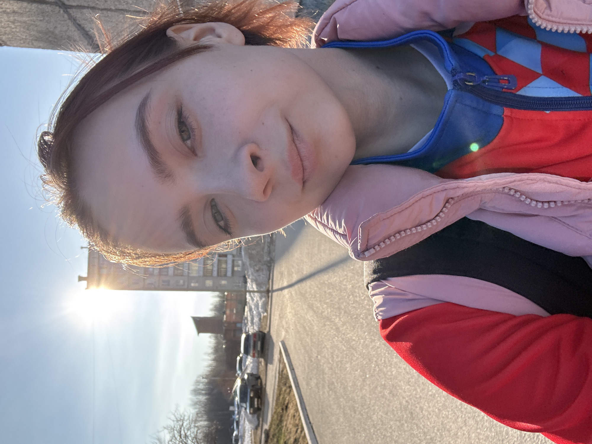 A person is pictured in a selfie in a sunny outdoor location. The person has fair skin, light eyes, and reddish-brown hair. They are wearing a jacket with a pink exterior, a blue inner lining, and red and blue checkered details on the chest. The person is slightly smiling.
The background shows an urban landscape. A building with multiple windows is visible in the distance, illuminated by the sun. There are parked cars along the side of a street. The sky is clear and blue. The scene is well-lit, indicating it is daytime. The location is city of Unknown, Unknown.
