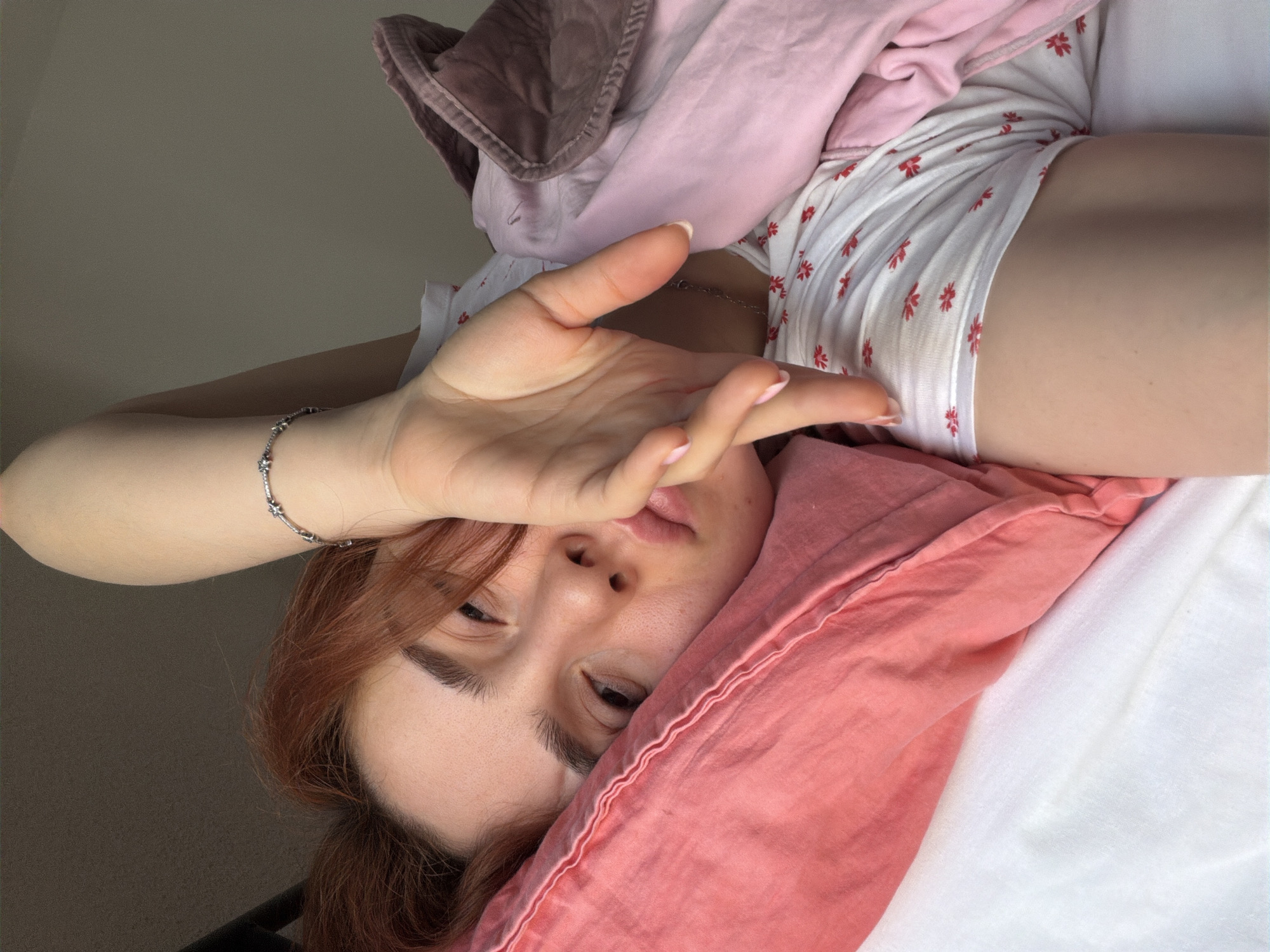 A young woman with auburn hair is lying in bed, propped up on a pink pillow. Her right arm is raised, with her hand covering part of her face. She is wearing a white t-shirt with a red floral pattern and a silver bracelet on her wrist. A pink blanket and a darker colored blanket are visible. The scene appears to be an indoor setting, possibly a bedroom. There is no indication of the location being in the city of Strasbourg, France.