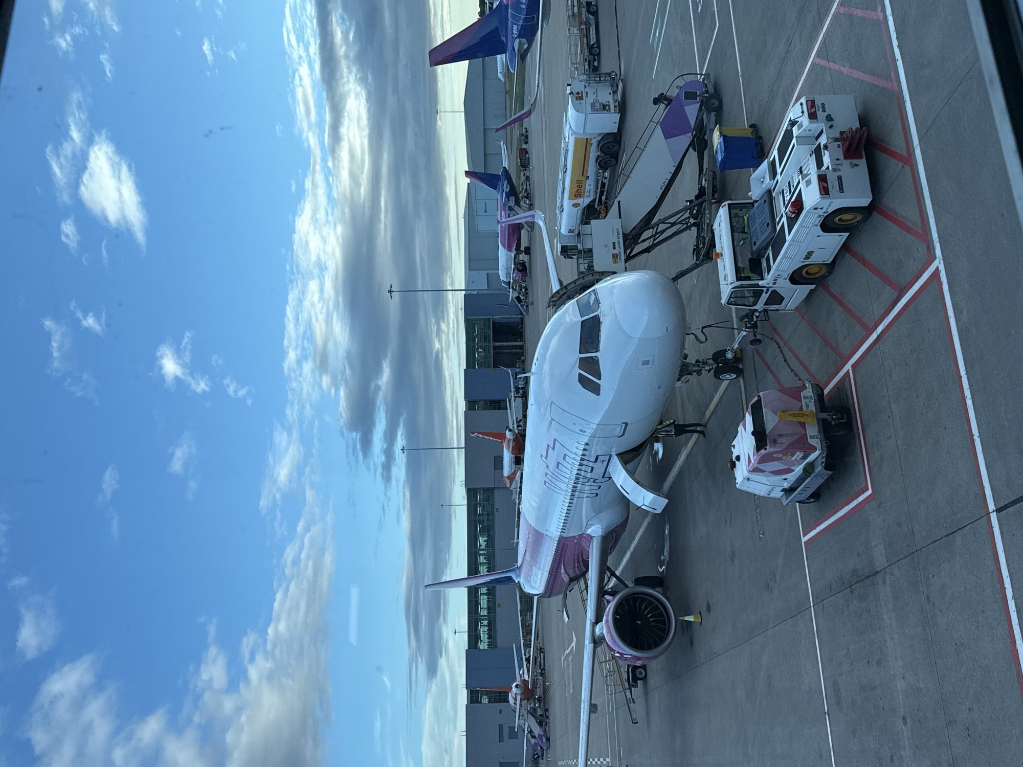 An aerial view of an airport tarmac shows multiple Wizz Air airplanes. In the foreground, a Wizz Air Airbus A320 with a pink tail and accents on its fuselage is parked. A tug vehicle is attached to the nose gear of the Airbus, and a fuel truck is positioned near the wing. In the middle ground, another Wizz Air Airbus A321 with a pink tail is parked. In the background, several other airplanes, including one with an orange tail, are visible. The airport terminal building is in the distance. The sky is partly cloudy.