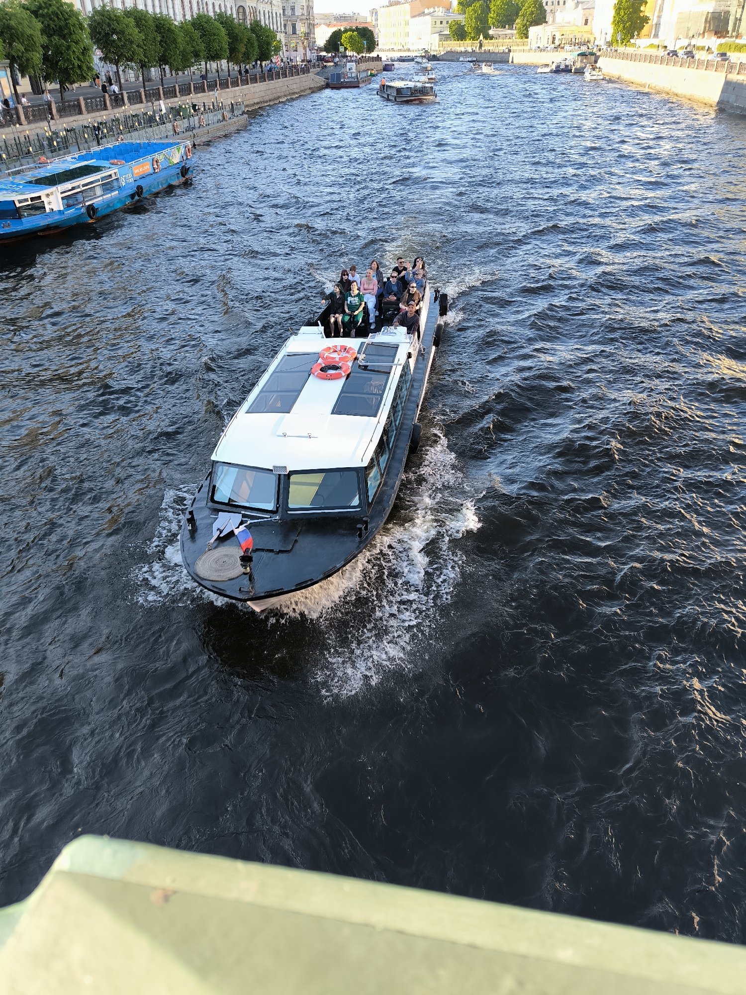 An overhead photograph captures a white and dark-colored passenger boat navigating a canal in Saint Petersburg, Russia, under clear daylight conditions.
In the foreground, a partial green metal bridge railing extends across the bottom left and right of the frame, indicating the vantage point. The central subject is a sleek, modern passenger boat, dark-colored below the waterline and white on its upper structure, moving from the middle right towards the upper left of the image. Its bow generates a distinct white spray and wake, while a smaller Russian flag (red, white, blue horizontal stripes) is visible on its foredeck, near a coiled rope. The boat's cabin features large, rectangular windows, and its white roof contains two transparent panels. Approximately 10 to 12 passengers are visible on the boat, primarily occupying the open-air stern section, where two orange life preservers are mounted on the white railing. Passengers are casually dressed, seated and standing, observing the surroundings. The boat's movement creates a prominent V-shaped wake trailing behind it on the dark blue-grey water, which is disturbed by numerous ripples.
The left bank of the canal features a stone embankment topped by a pedestrian walkway. A continuous row of mature green trees borders the walkway, separating it from a large, multi-story historical building with classical architectural elements and numerous windows. Adjacent to this embankment, in the mid-ground, a long, blue passenger boat is docked, displaying "СПБ" (Saint Petersburg) and other Cyrillic text on its side, confirming the location. Several orange life preservers are also visible along the blue boat's hull. Indistinct figures of people are visible walking along the embankment in the distance.
The right bank, receding into the background, also features stone embankments and light-colored historical buildings. Several other smaller boats are visible in the far distance, navigating or docked on the canal. The overall scene is brightly lit by natural sunlight, suggesting a clear day.